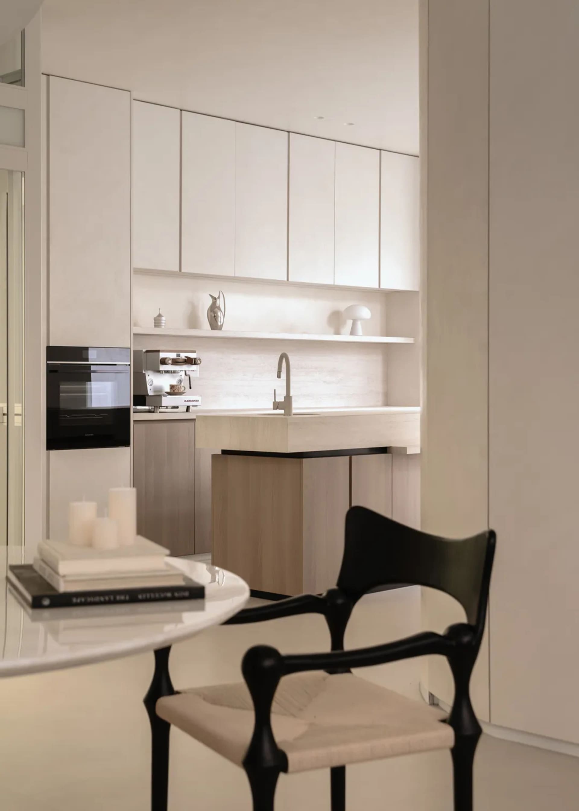 Modern minimalist kitchen with white matte cabinetry, light wood breakfast bar, built-in oven, open shelving, and a sculptural black chair in the foreground.