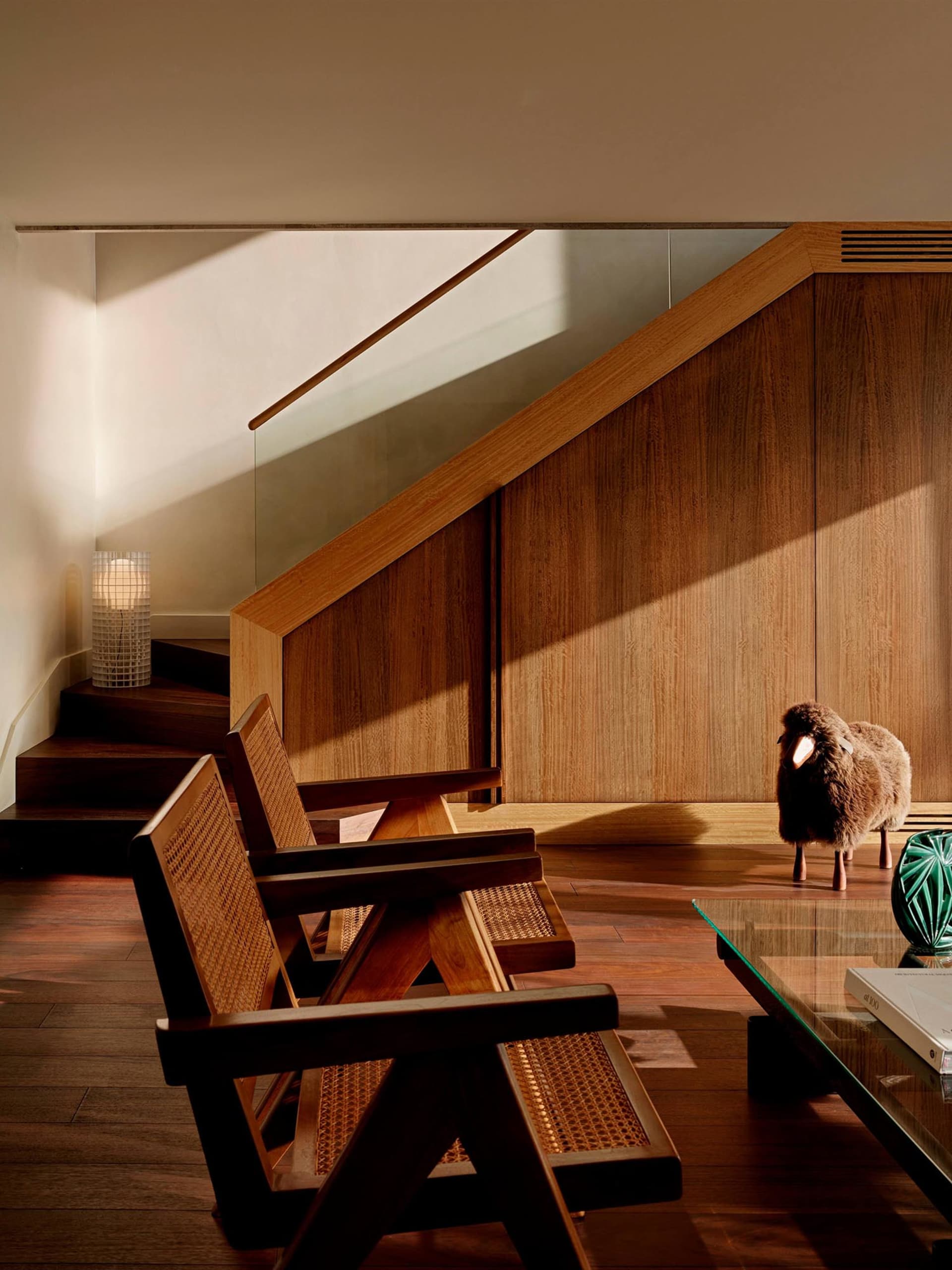 Two wooden armchairs with cane backs placed under a wooden staircase, next to a glass coffee table and a wool sheep sculpture in a sunlit modern interior.