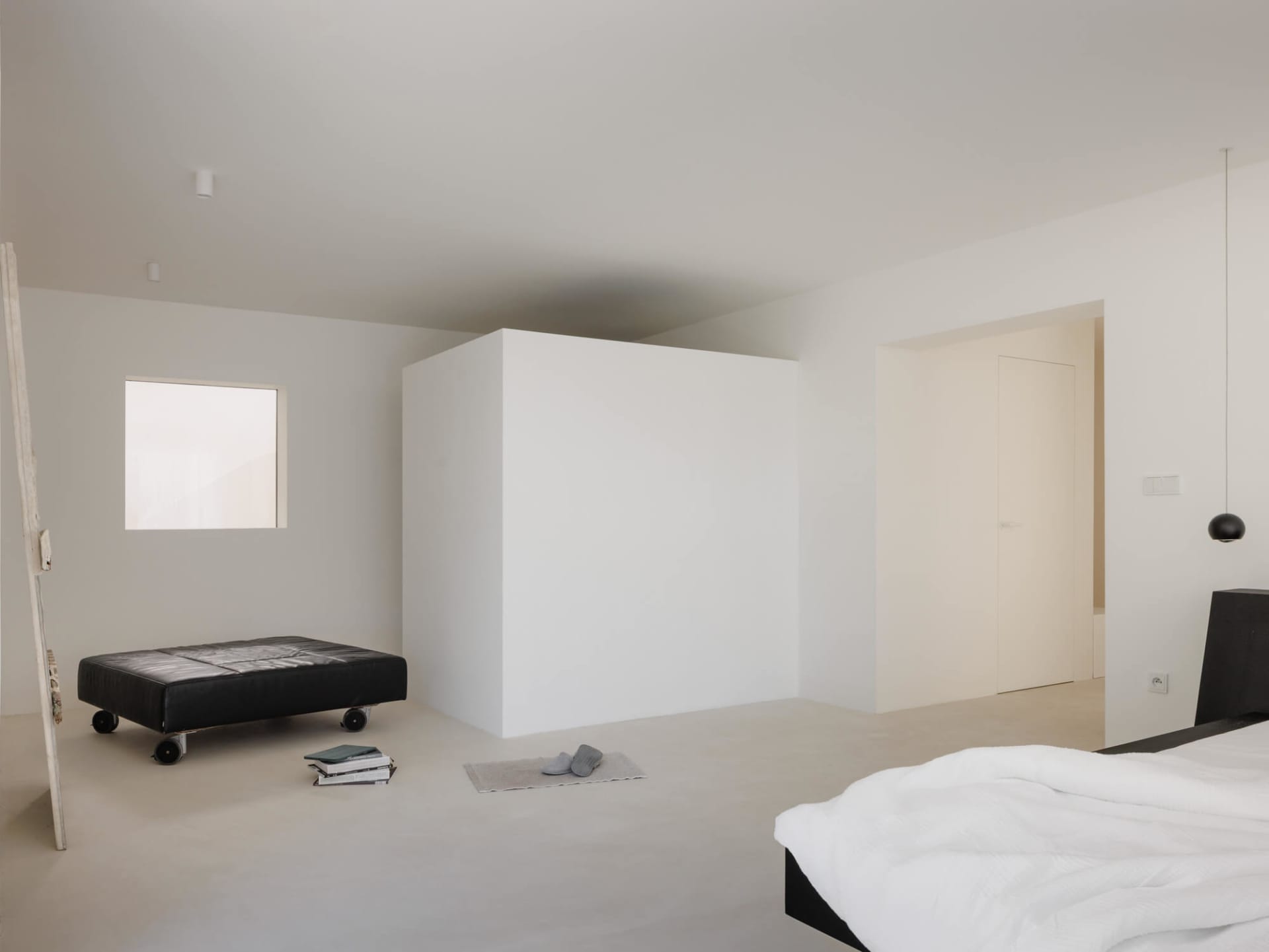 Minimalist bedroom interior with white walls, polished concrete floor, black low platform bed with white bedding, black leather rolling ottoman, and a small square window providing natural light.