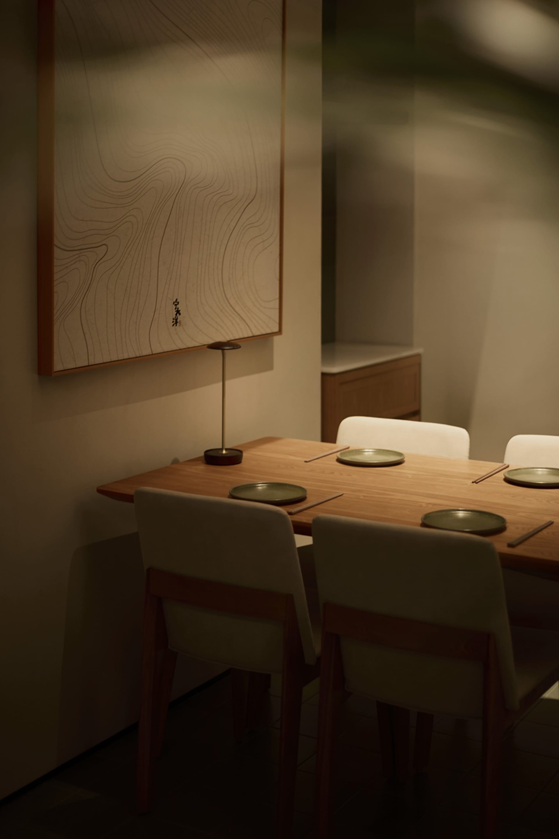 Modern minimalist dining room featuring a solid oak table, four upholstered chairs with wooden frames, a sculptural table lamp, and a large framed Japanese line-drawing wall art with calligraphy.