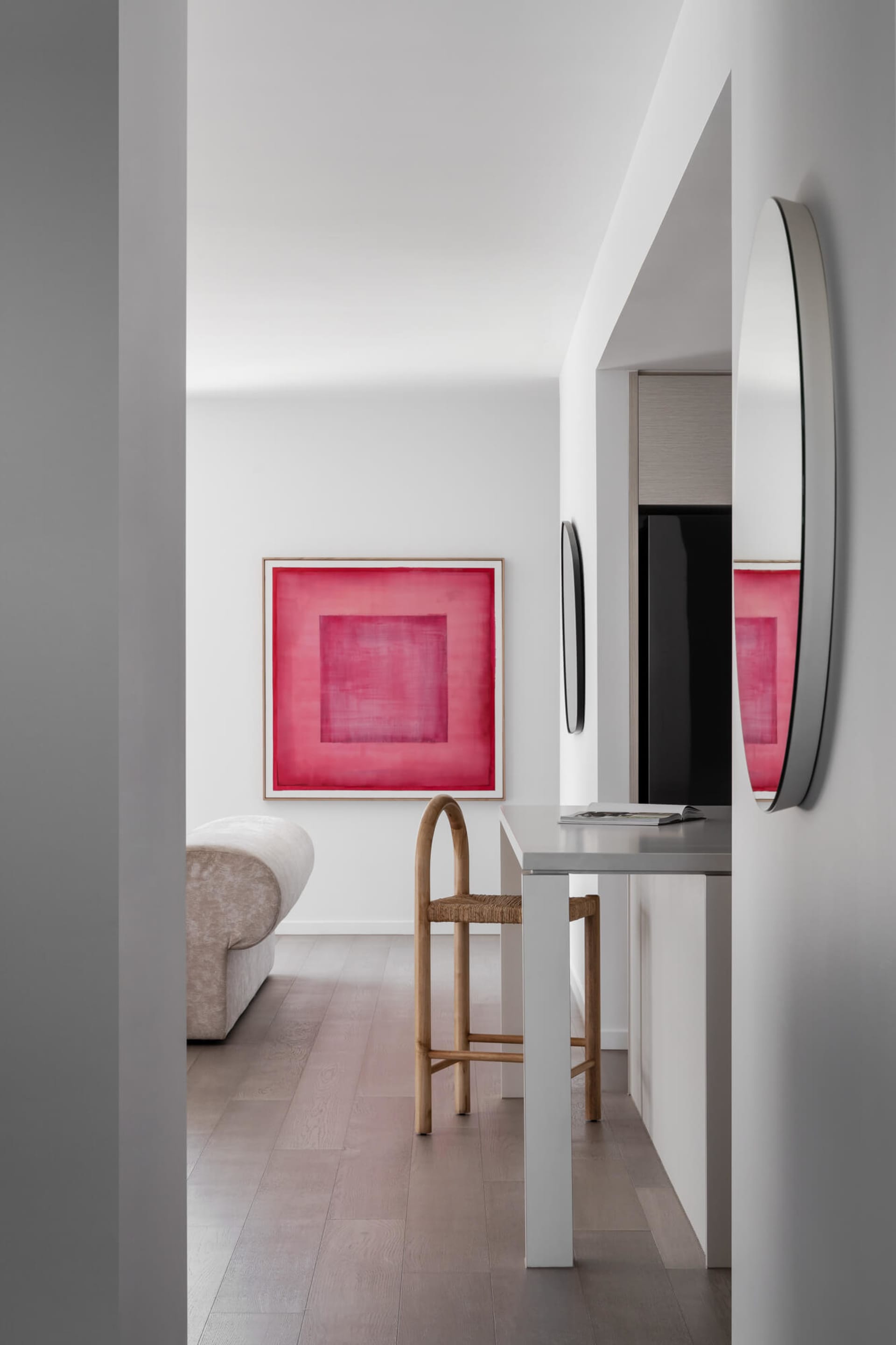 Modern minimalist entryway with white walls, light oak flooring, a white console table paired with a bentwood and rattan stool, and a large framed pink abstract painting on the back wall.