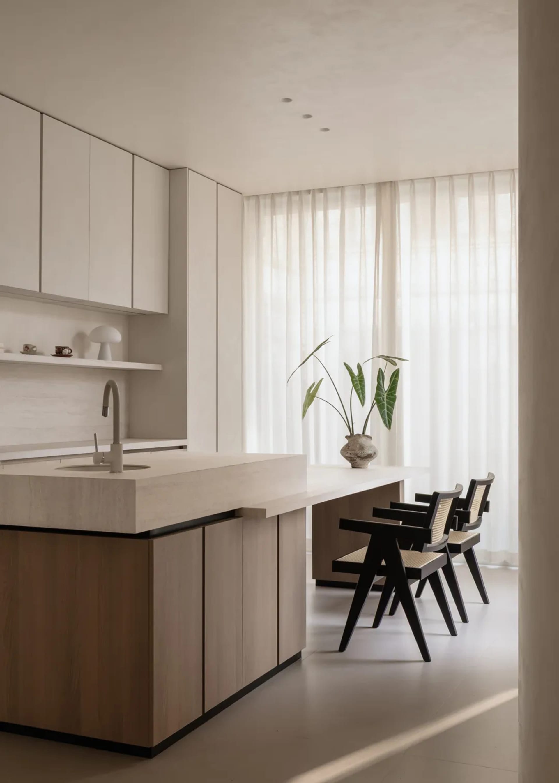 Modern minimalist kitchen featuring a large oak island with integrated sink, white upper cabinets, and three black dining chairs with cane backs placed beside a long table near a window with sheer curtains.