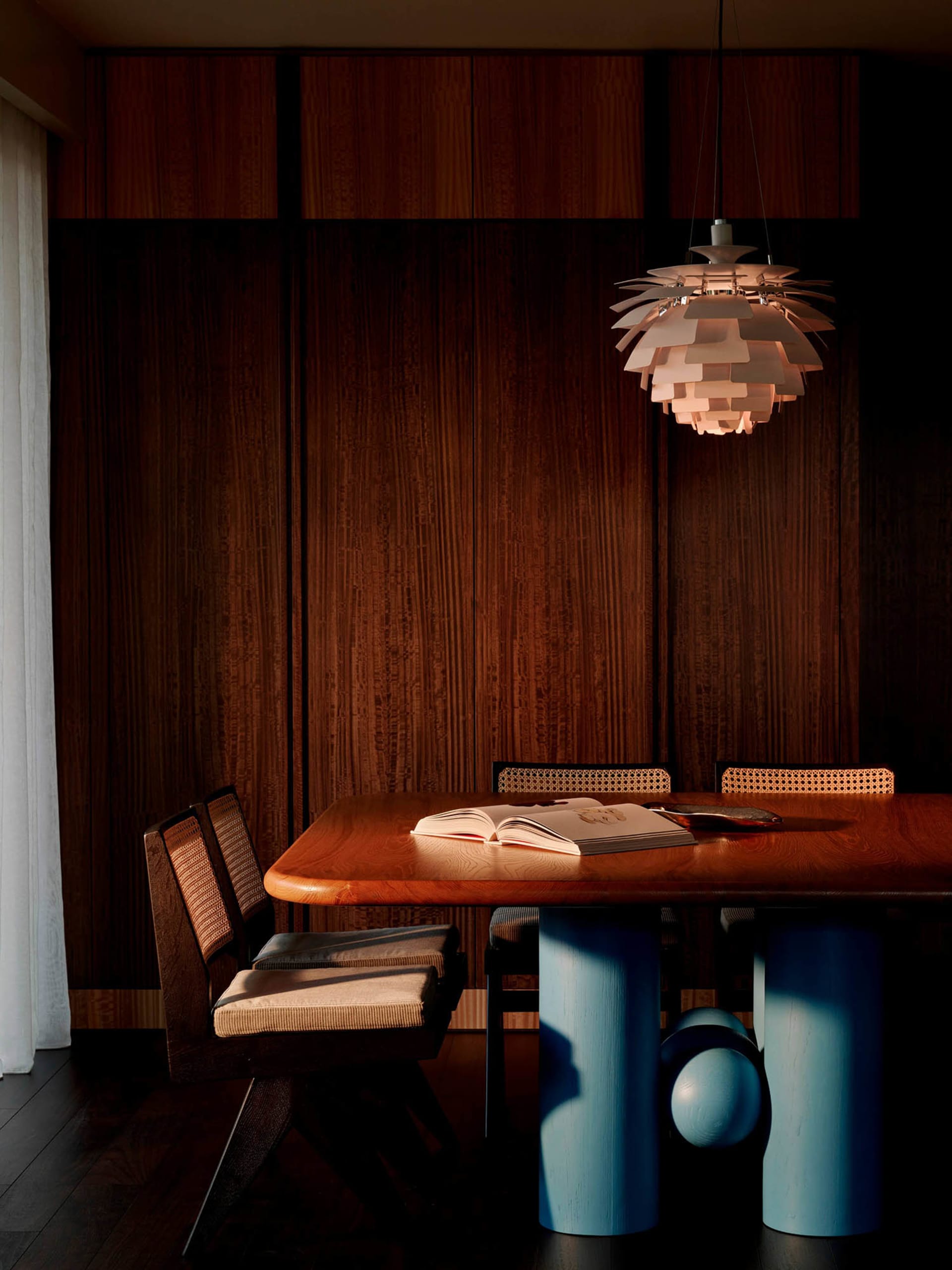 Modern dining room featuring a walnut dining table with bright blue cylindrical pedestal legs, cane-back dining chairs, and a large white Artichoke pendant lamp hanging above an open book on the table.
