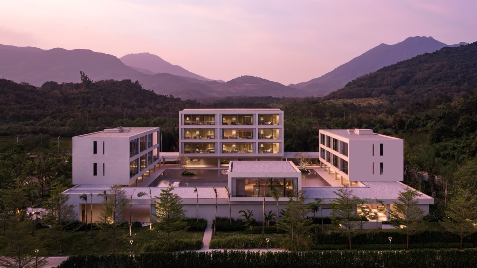 Aerial view of a contemporary concrete resort complex with illuminated interiors, surrounded by tropical landscaping and dramatic mountains at twilight.