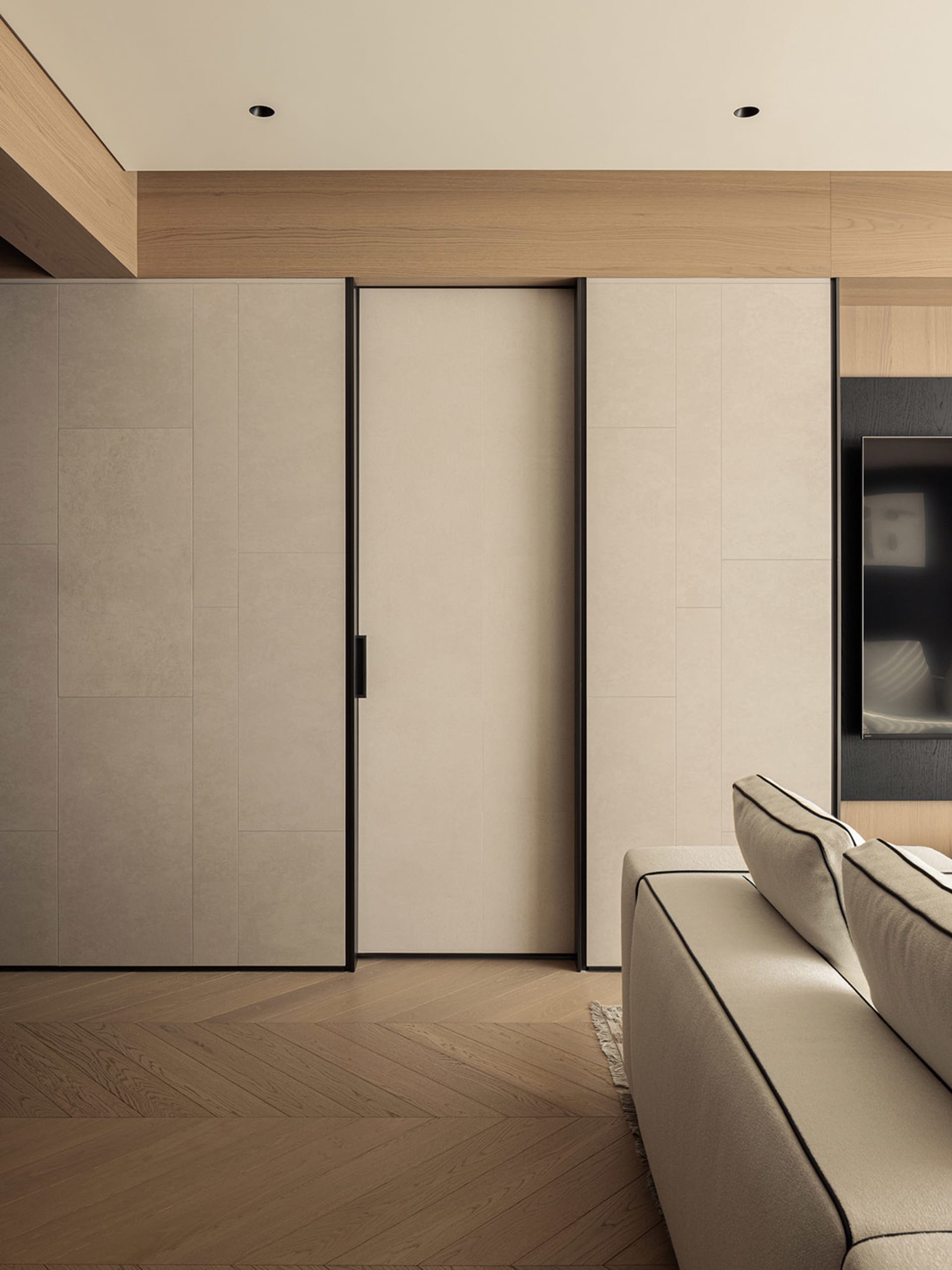 Modern minimalist living room featuring beige built-in cabinetry with black sliding door, light oak herringbone wood floor, and a portion of a neutral sofa with black piping.