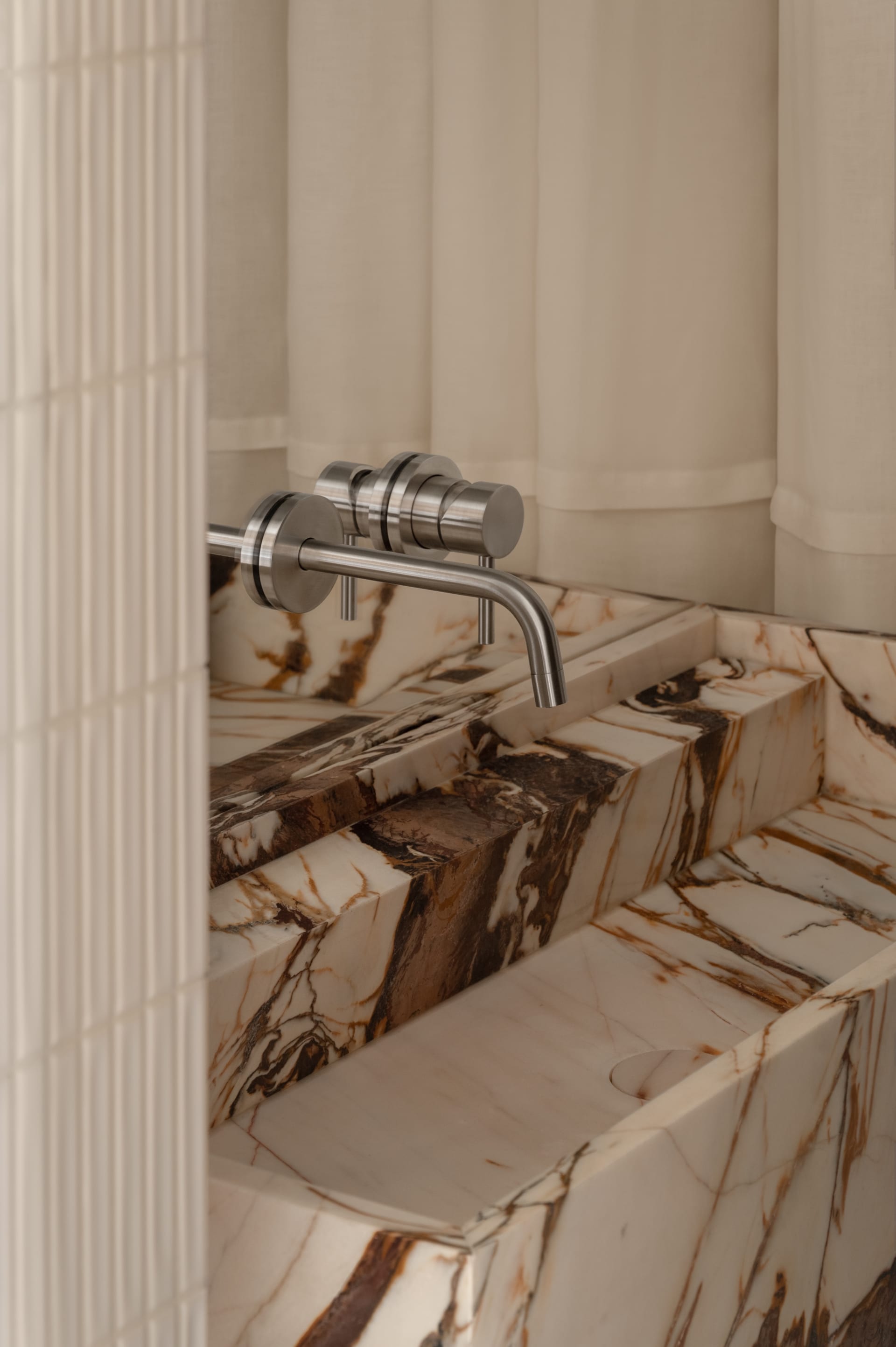Close-up of a luxurious Calacatta marble vanity sink with dramatic brown veining and a modern brushed nickel wall-mounted faucet against a neutral textured wall.