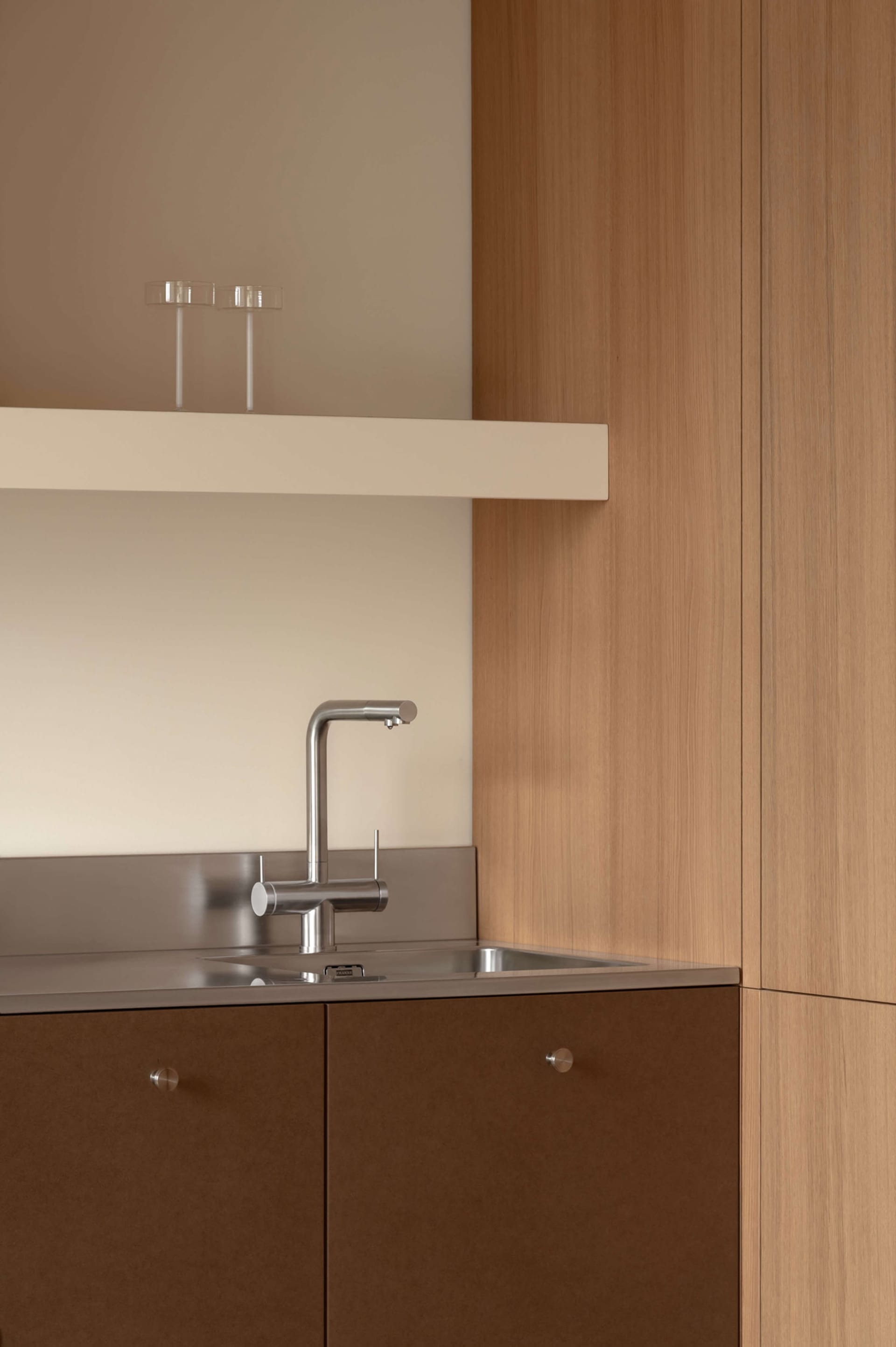 Close-up of a modern kitchen sink area with stainless steel faucet, integrated stainless steel backsplash and sink, warm wood lower cabinets with round pulls, and a floating white shelf above with two glass tumblers.