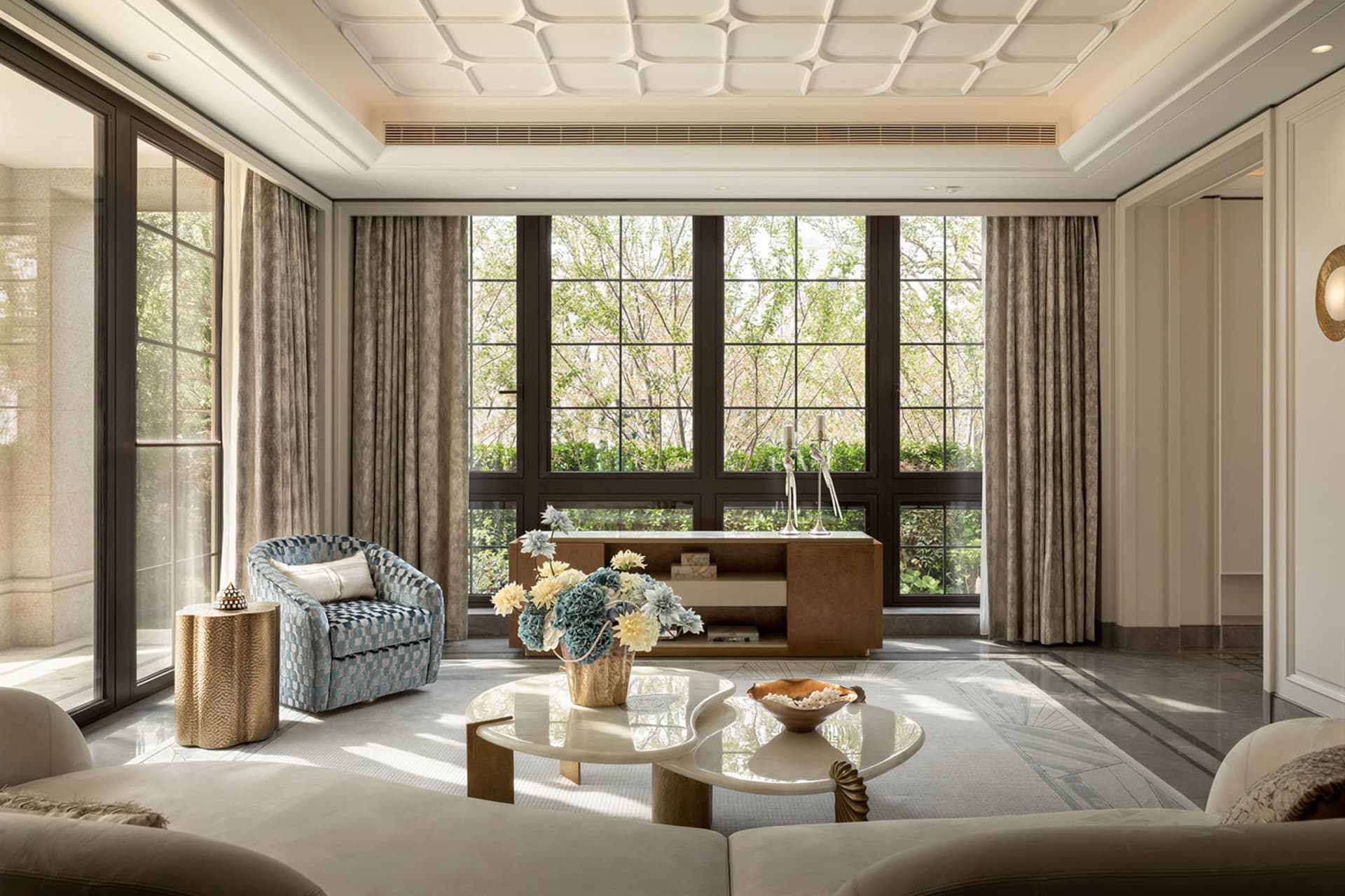 Bright modern luxury living room with large windows overlooking greenery, featuring a blue and white patterned armchair, gold textured side table, sculptural white marble coffee table, and neutral sofa.