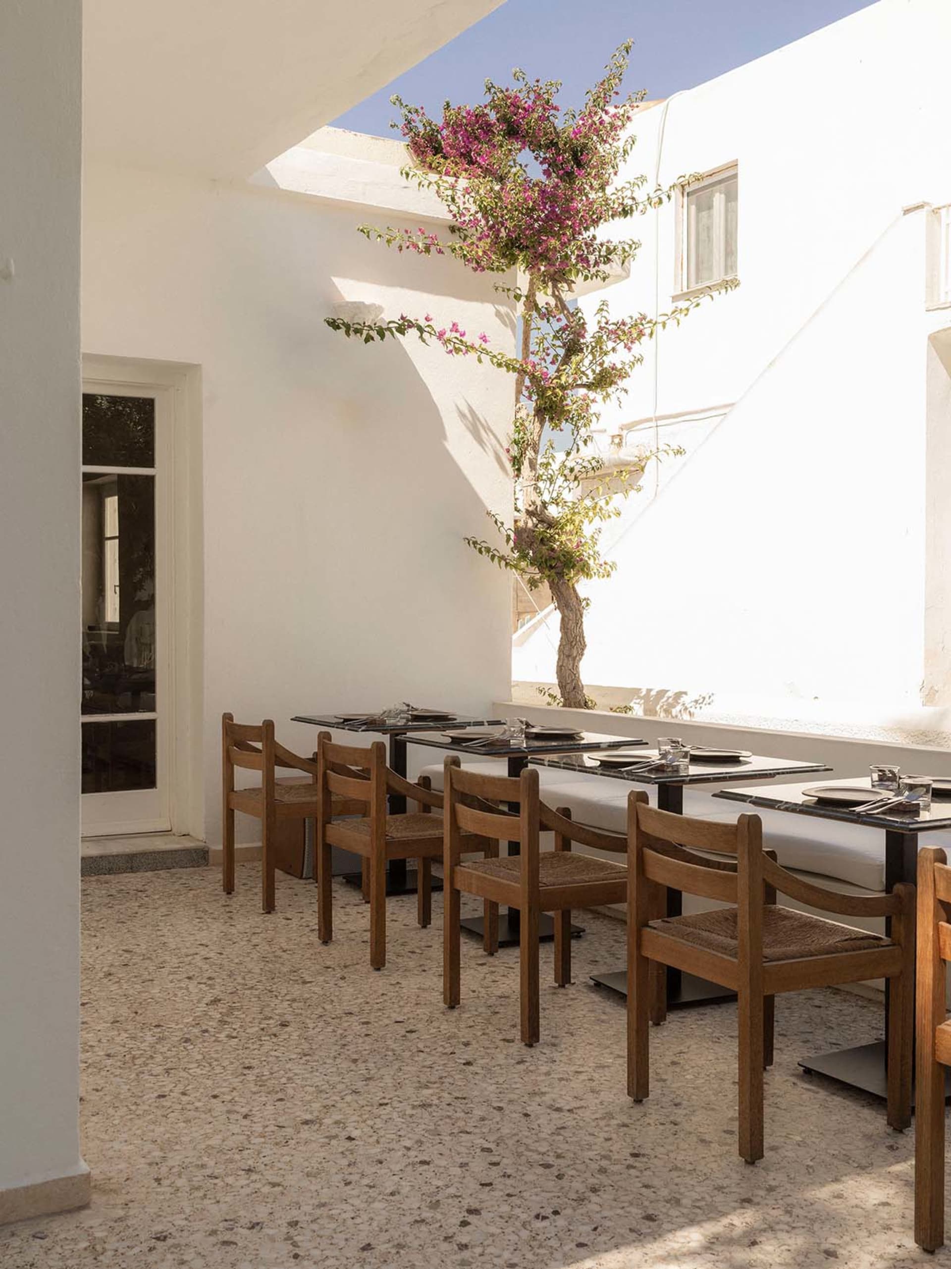 Outdoor courtyard dining area with wooden chairs, black marble tables, terrazzo floor, and a blooming bougainvillea tree under a bright blue sky.
