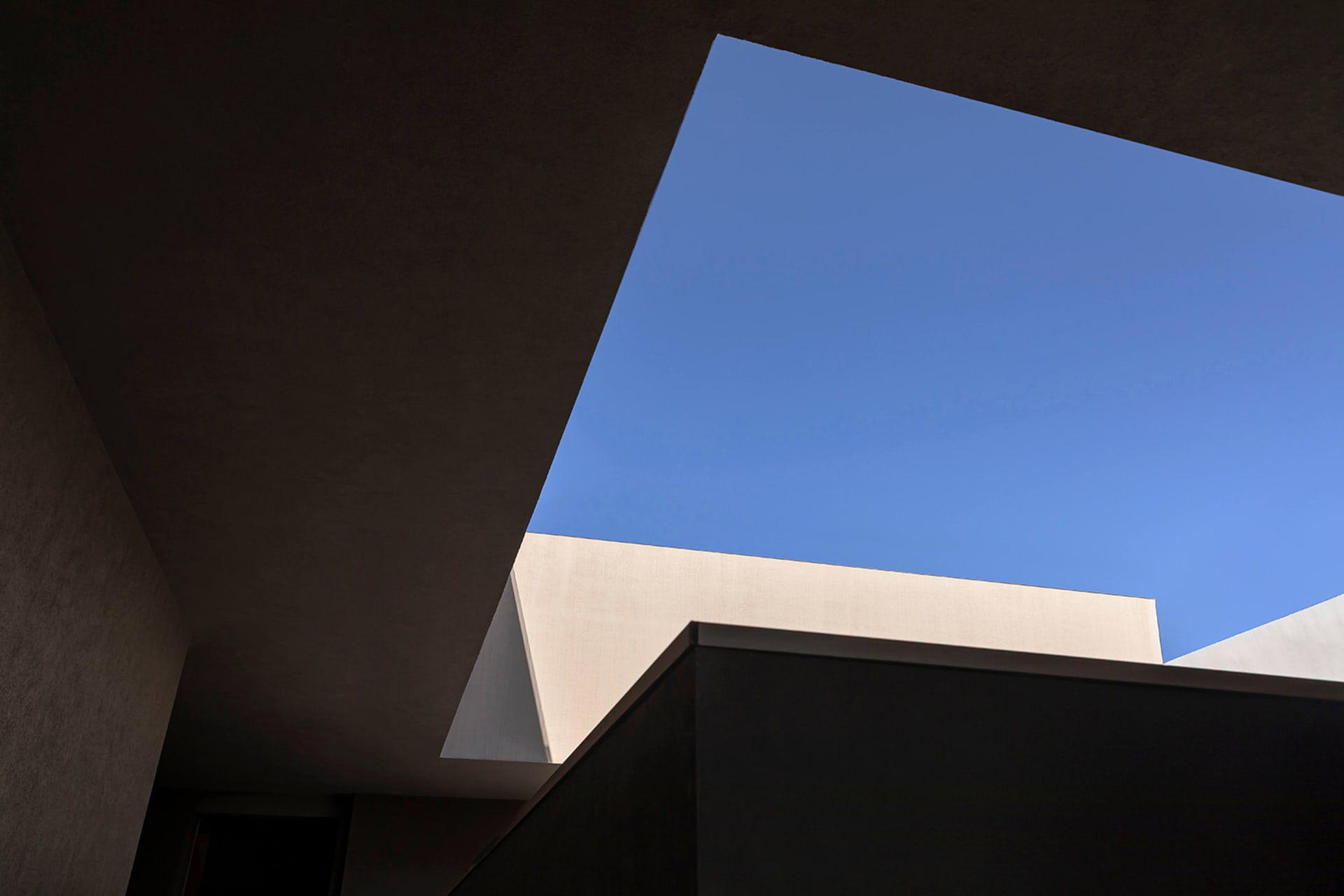 Low-angle architectural view looking up through a concrete building structure toward a clear blue sky, showing minimalist geometric forms and material textures.