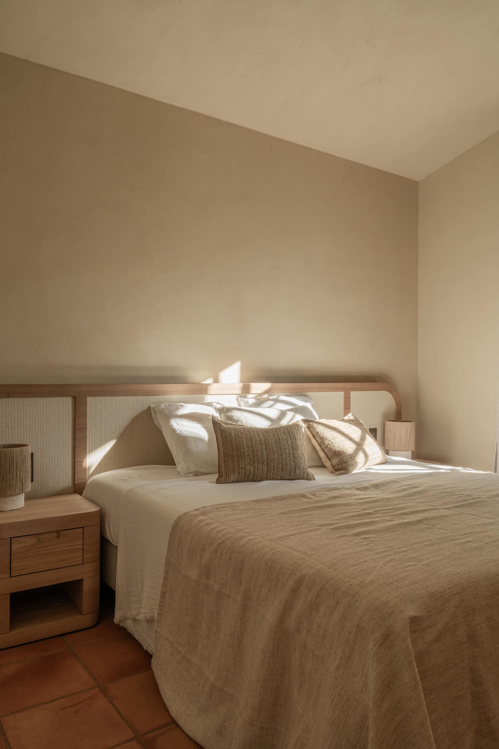 A minimalist bedroom featuring a low wooden bed with textured beige linens, terracotta floor tiles, and warm sunlight streaming through a window creating strong diagonal shadows on the wall and bedding.