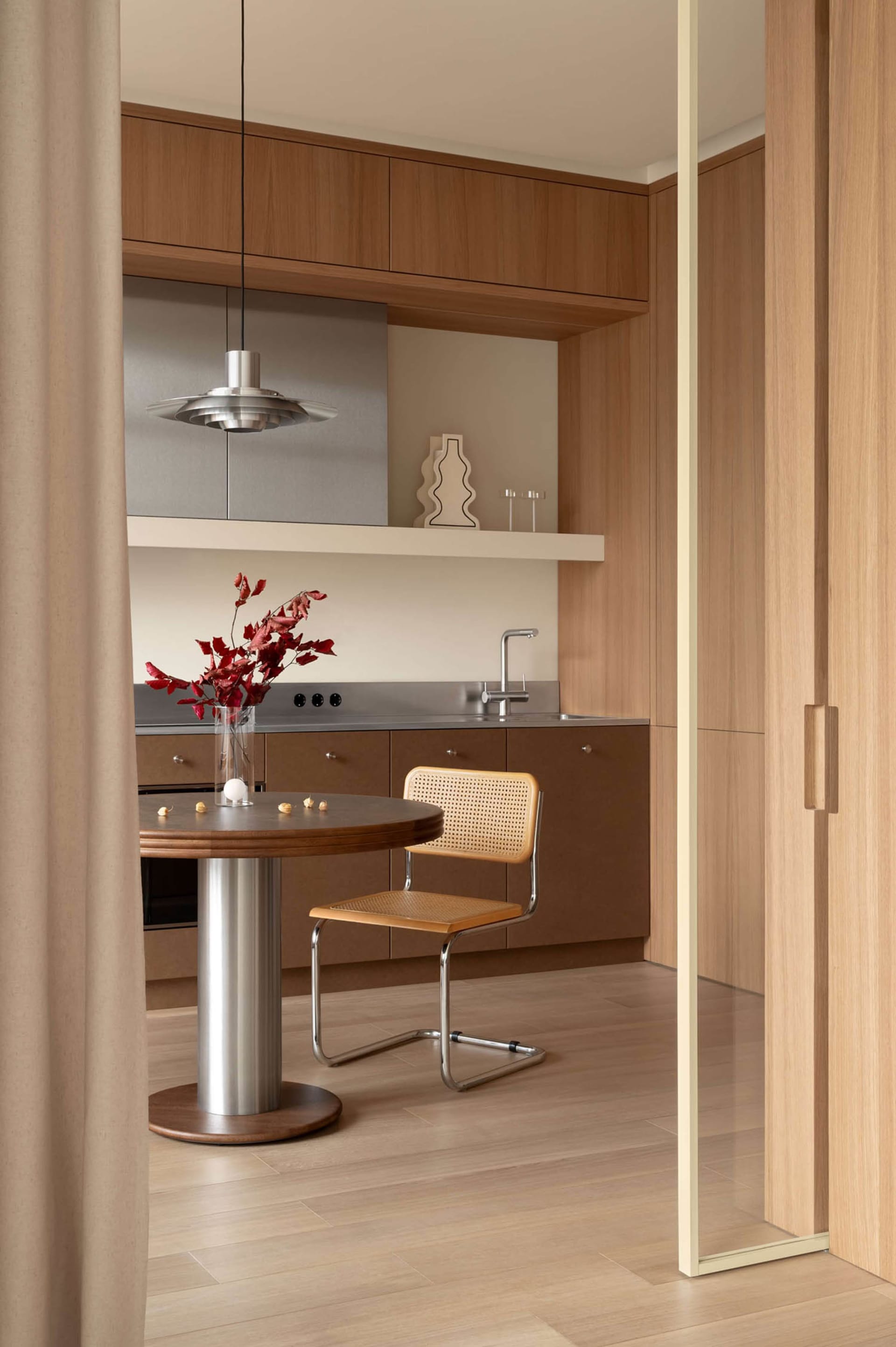 View of a modern minimalist kitchen dining area with walnut cabinetry, a round wooden dining table on a metal pedestal, a cane and leather Cesca-style chair, stainless steel countertop and sink, and a large metallic pendant lamp.