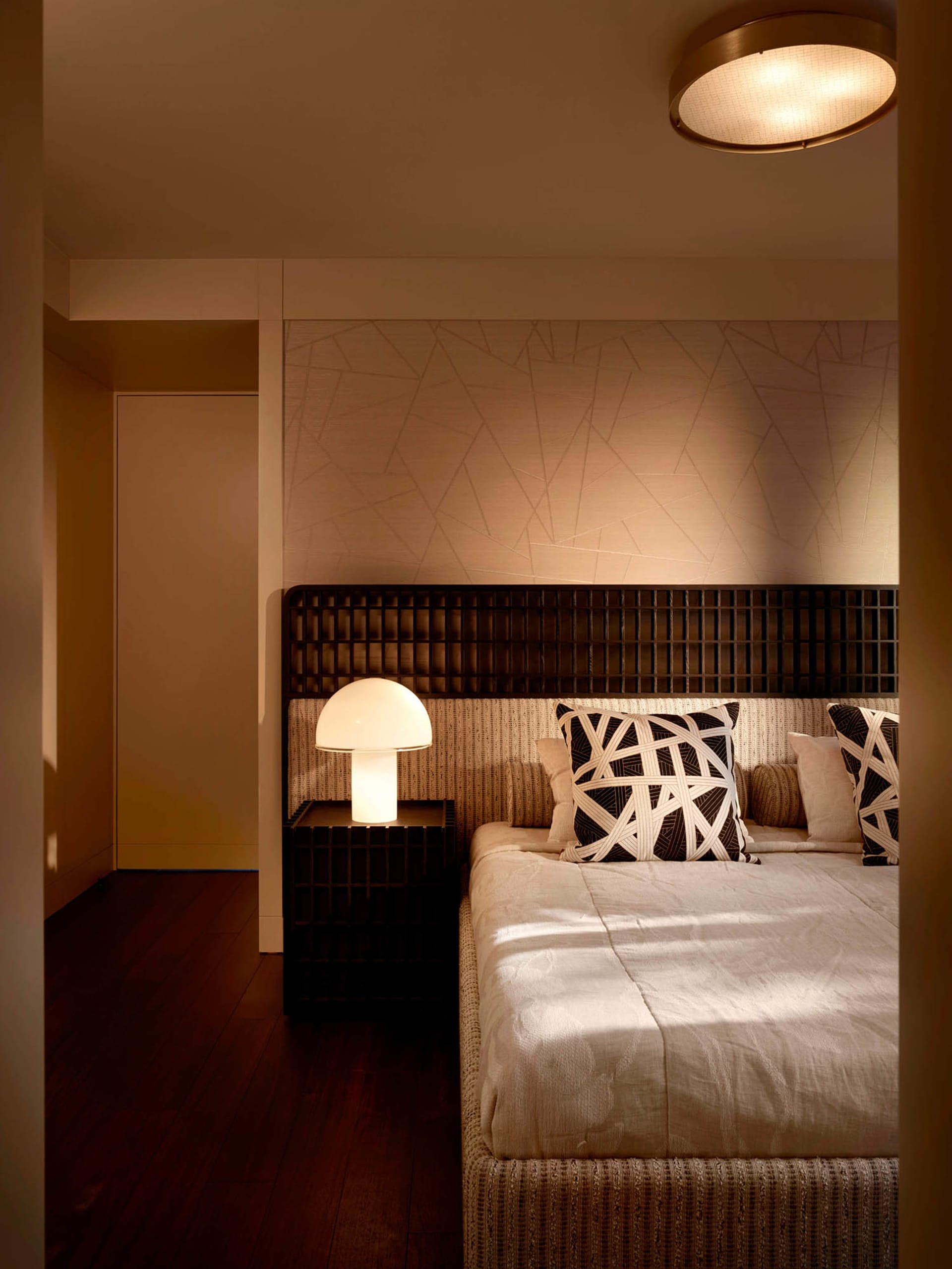 Modern minimalist bedroom with dark wood grid headboard, textured beige wallpaper with geometric line pattern, white mushroom table lamp, and black and white abstract pillows on a woven bed base.