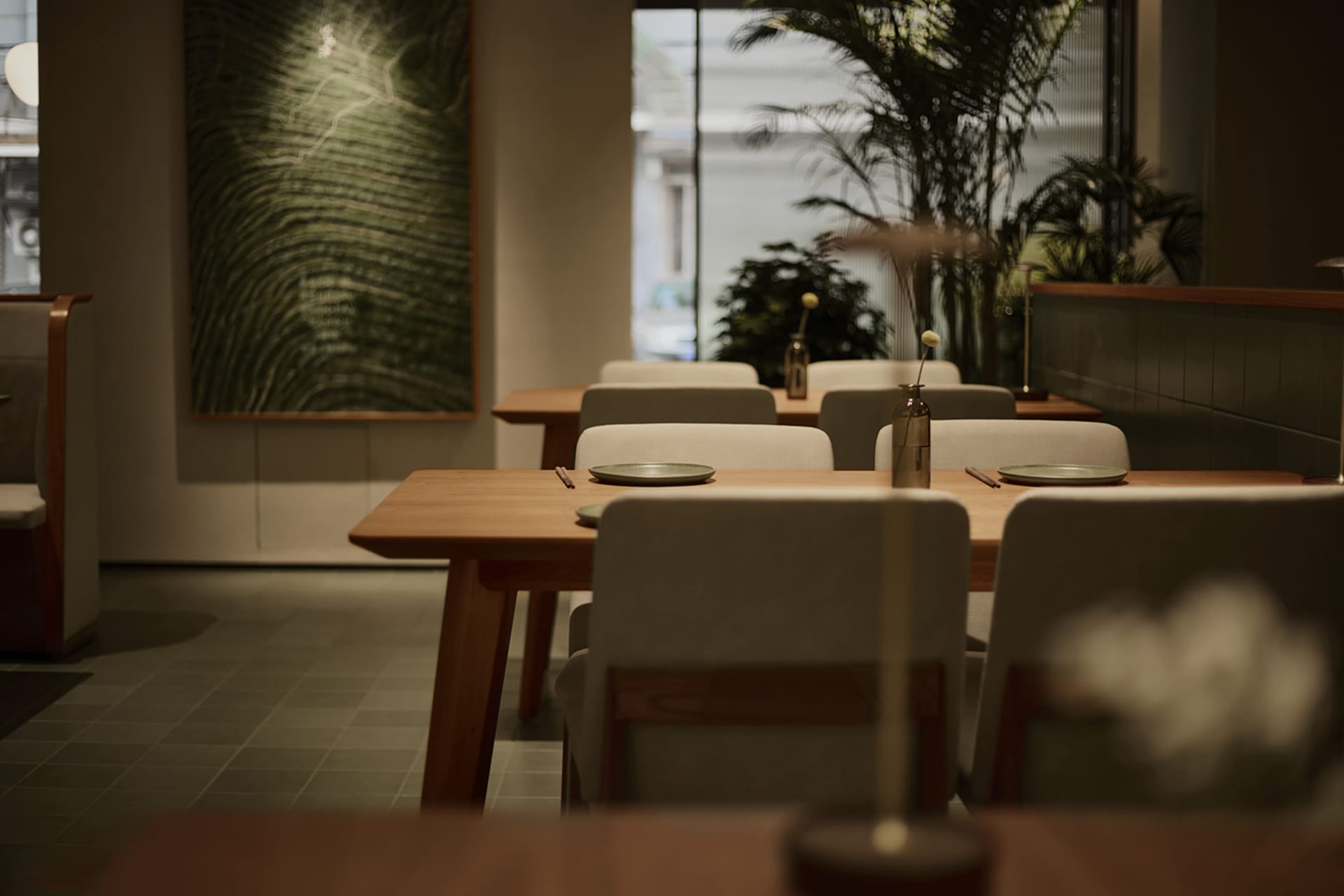 Modern minimalist restaurant dining area featuring light oak wooden tables, beige upholstered chairs, large textured green artwork on the wall, and abundant indoor plants near large windows.
