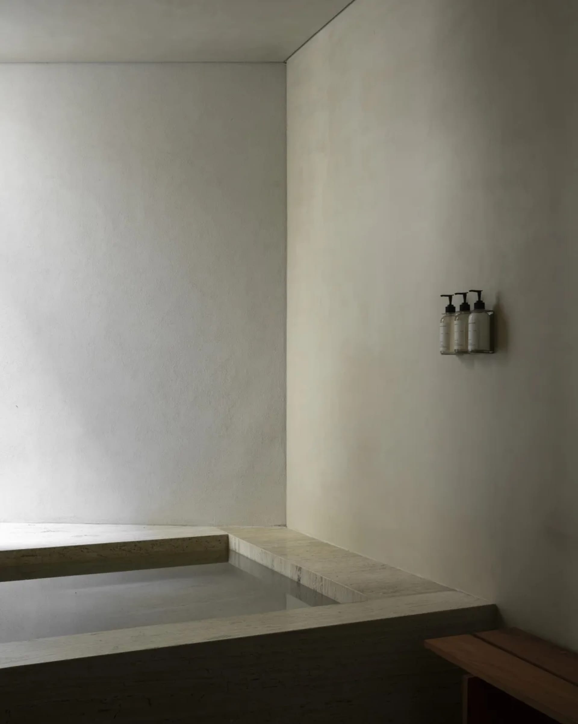 Minimalist bathroom corner featuring a sunken travertine bathtub with integrated stone ledge and wall-mounted soap dispensers.