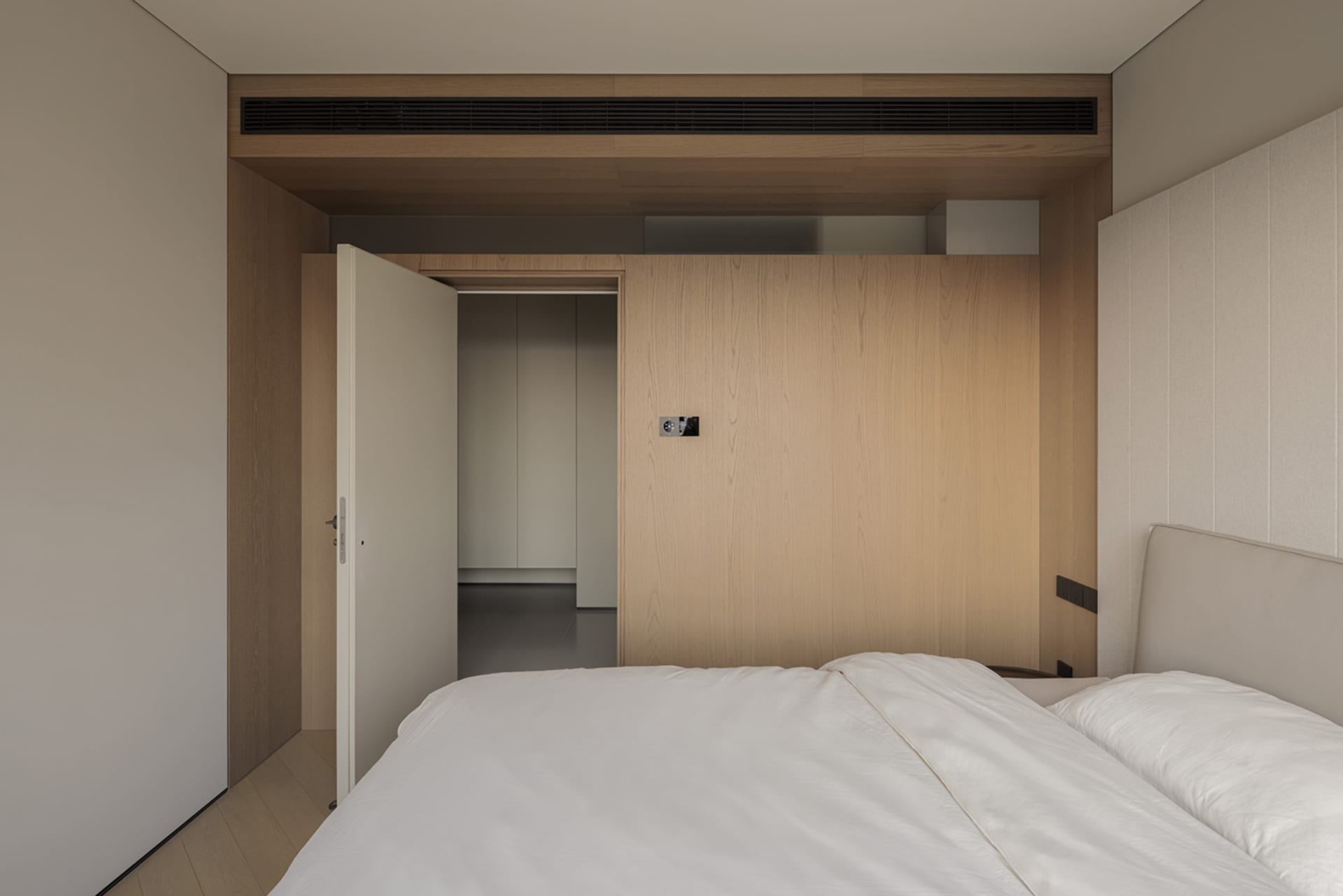 Modern minimalist bedroom with a large light oak built-in wardrobe, upholstered headboard, white bedding, and an open doorway leading to a hallway.