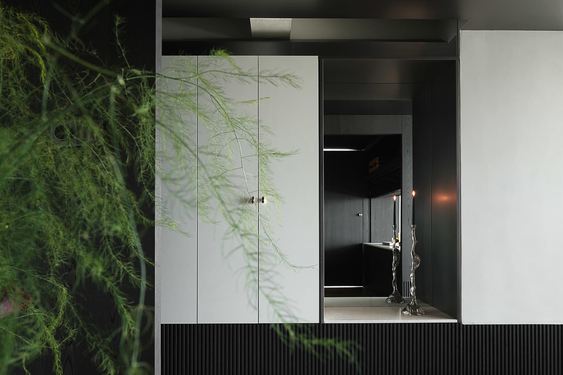Modern dark entryway featuring white matte lacquer built-in cabinet, black ribbed paneling, two lit sculptural candlesticks, and a large hanging asparagus fern in the foreground.