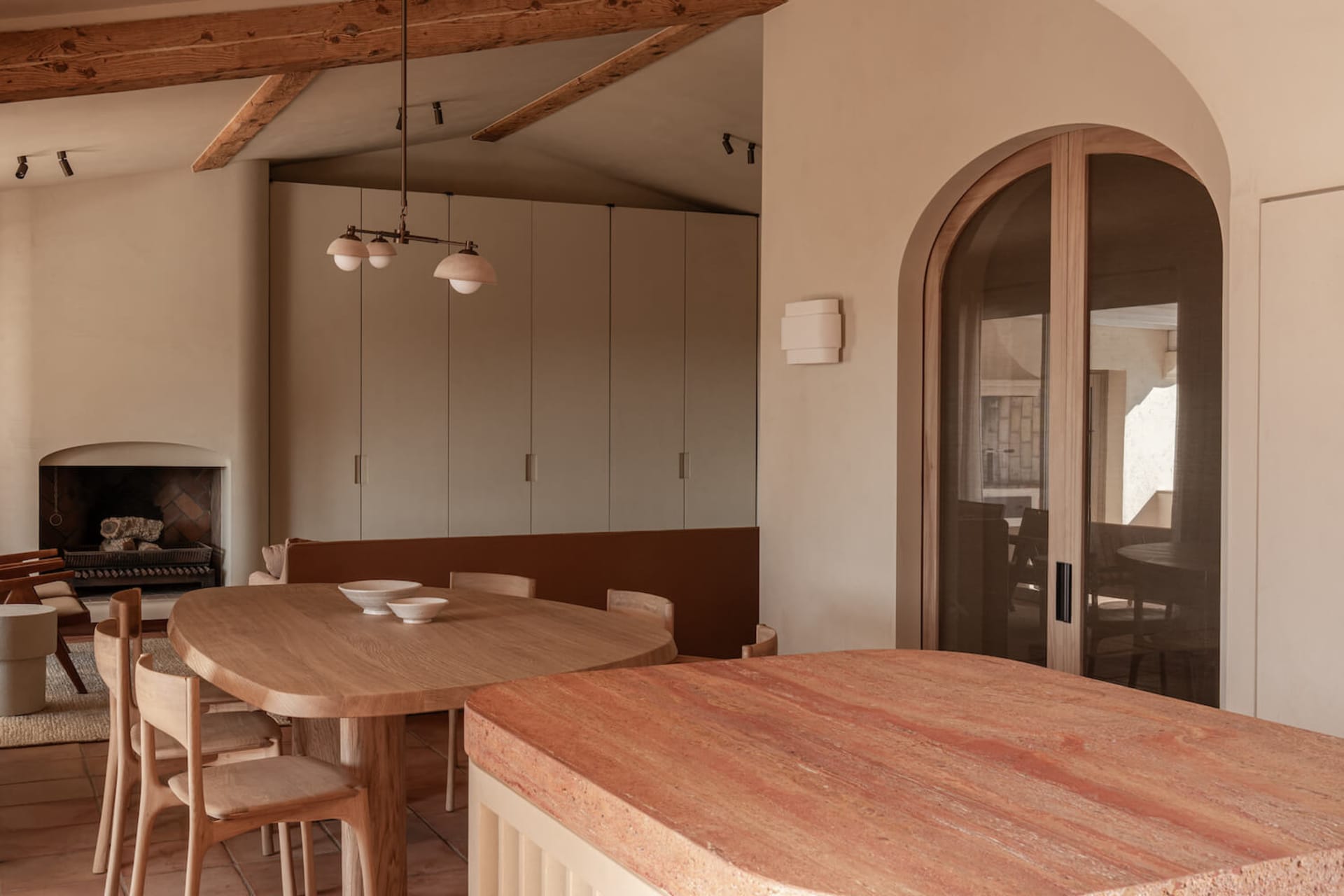 Modern minimalist dining room featuring a large oval oak dining table, wooden chairs, a terracotta stone kitchen island in the foreground, built-in banquette seating, exposed wooden ceiling beams, and a fireplace in the background.