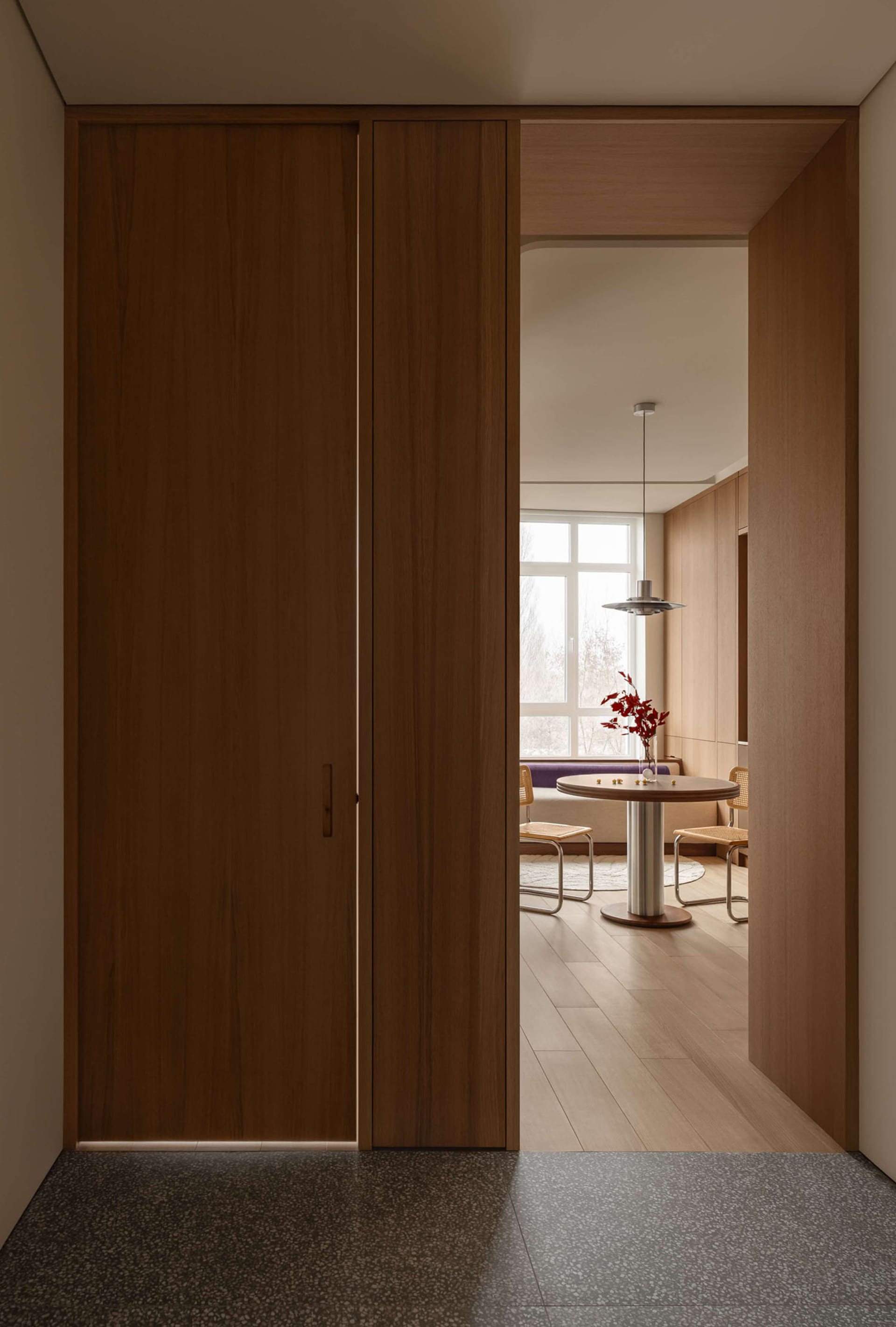 Modern minimalist interior showing a tall walnut wood pivot door partially open, revealing a bright dining area with round wooden table, woven chairs, pendant light, and window seat in the background.