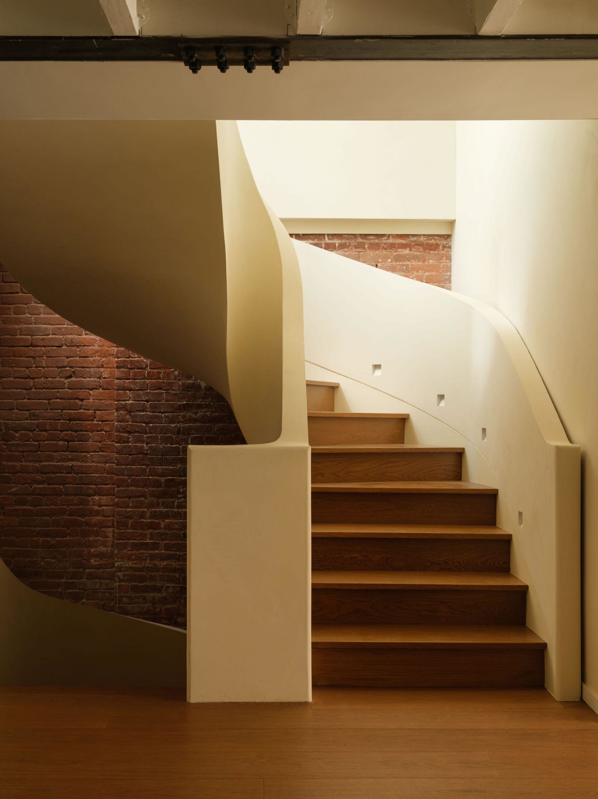 View of a modern curved wooden staircase with light-colored plaster walls and an exposed red brick wall in the background.