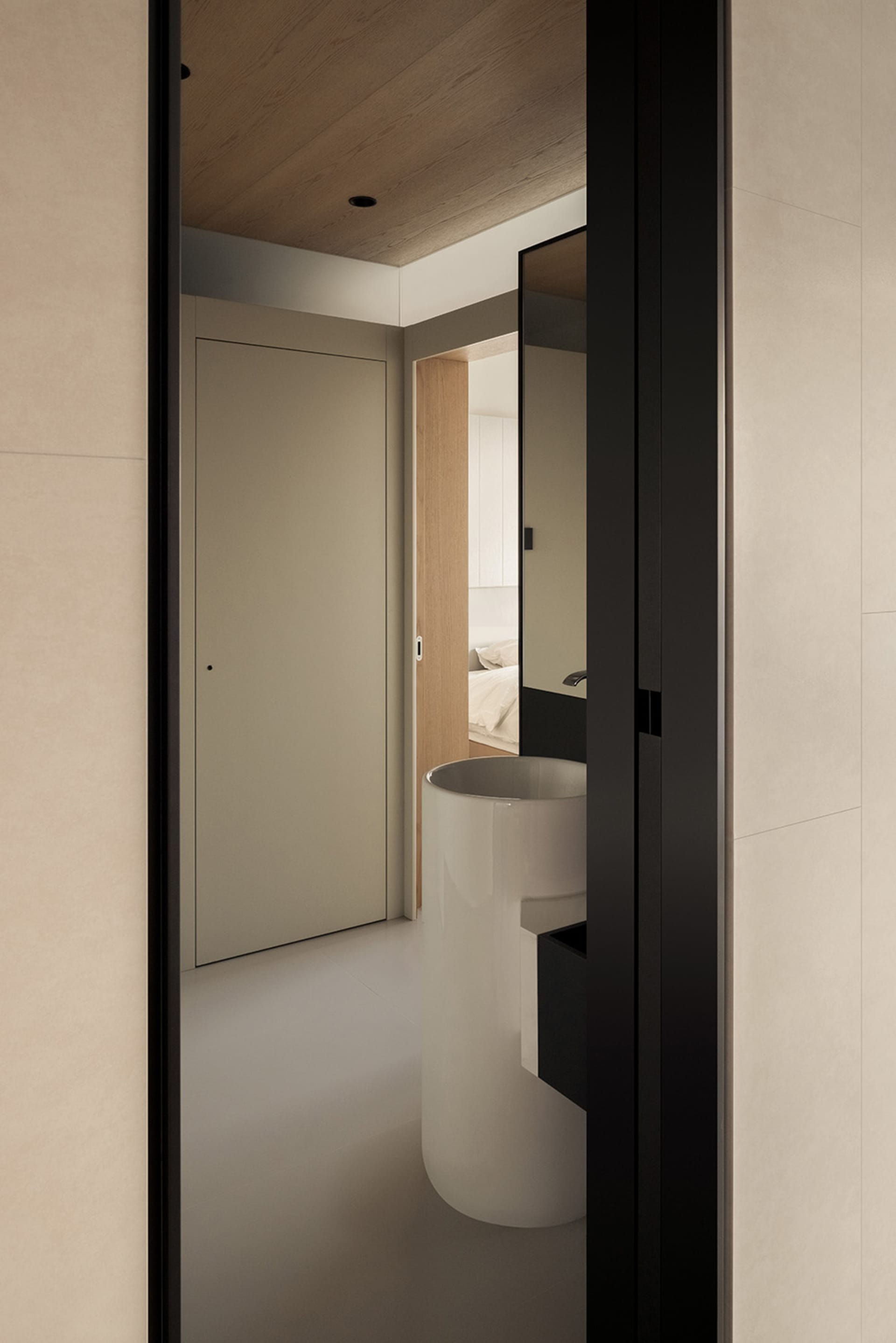 Modern minimalist bathroom featuring a cylindrical white porcelain sink with black floating vanity, viewed through a dark door frame with wood ceiling and neutral walls.