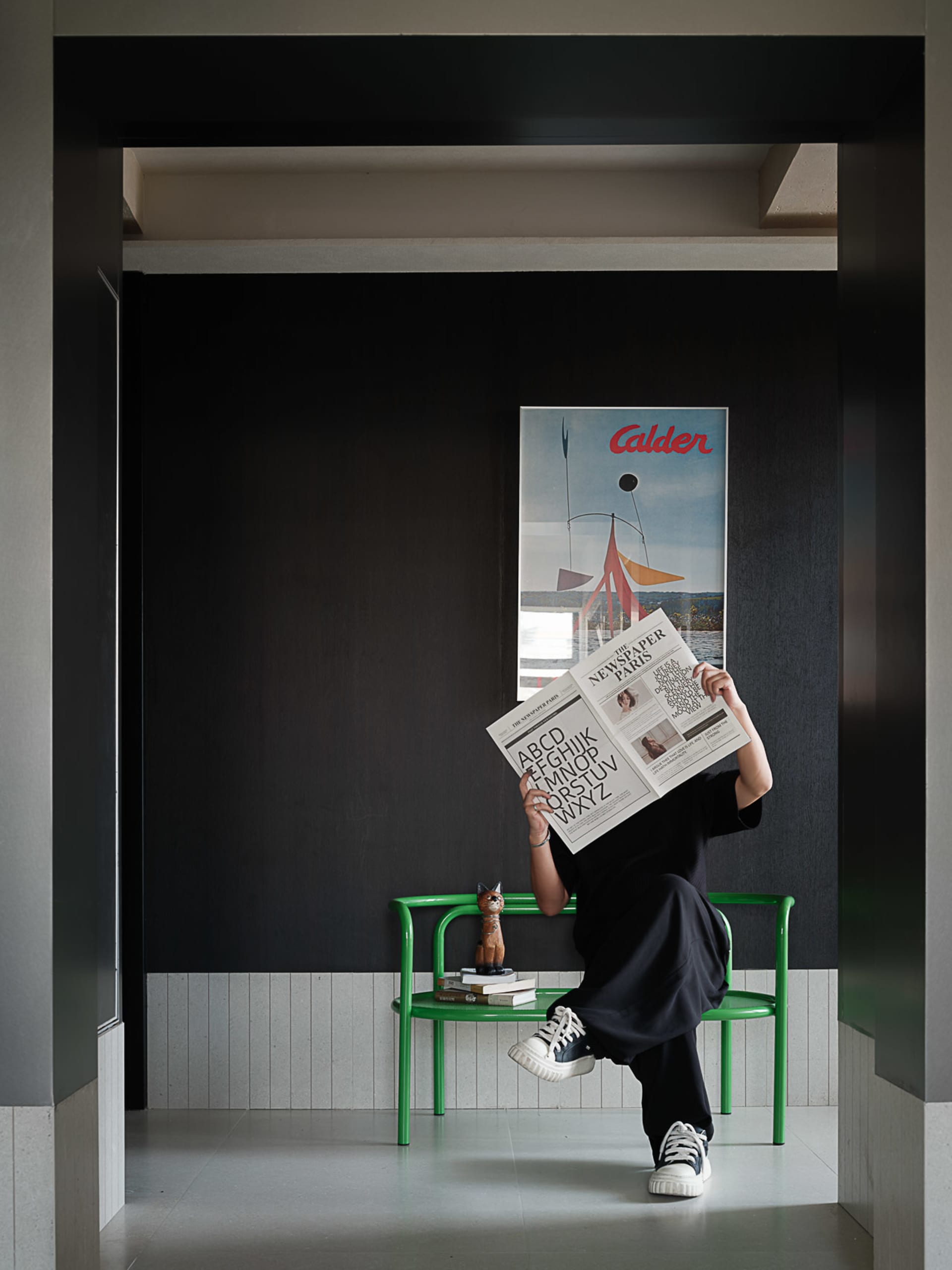 Person reading a newspaper while sitting on a bright green chair in a modern dark entryway with a large Alexander Calder poster on the wall.