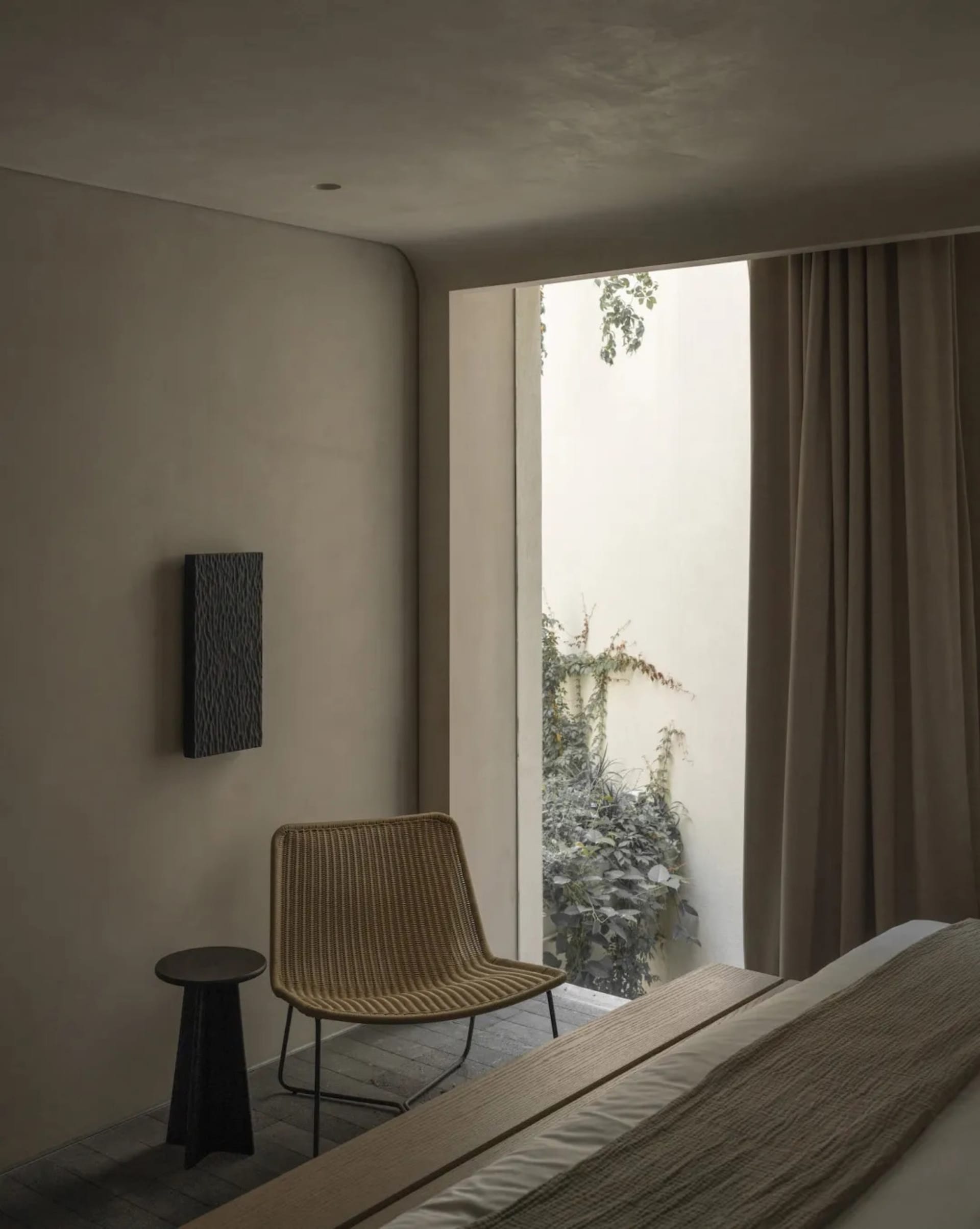 Minimalist bedroom interior featuring a woven rattan chair, dark side table, textured wall art, and large opening to a lush green courtyard with beige curtains.