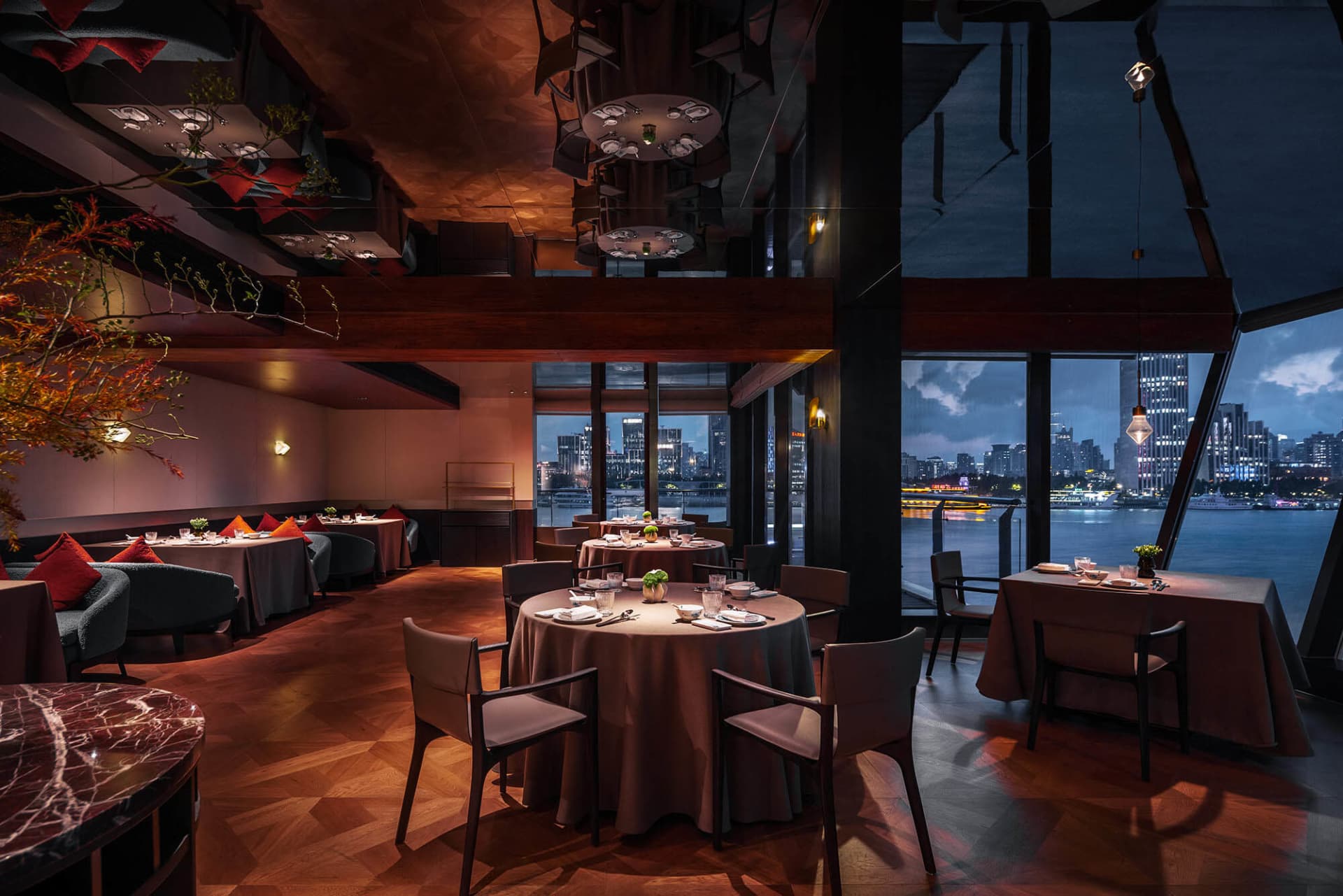 Luxurious modern restaurant interior featuring dark walnut wood ceiling, geometric parquet floor, round dining tables with taupe linens, and large windows overlooking a river and illuminated city skyline at dusk.