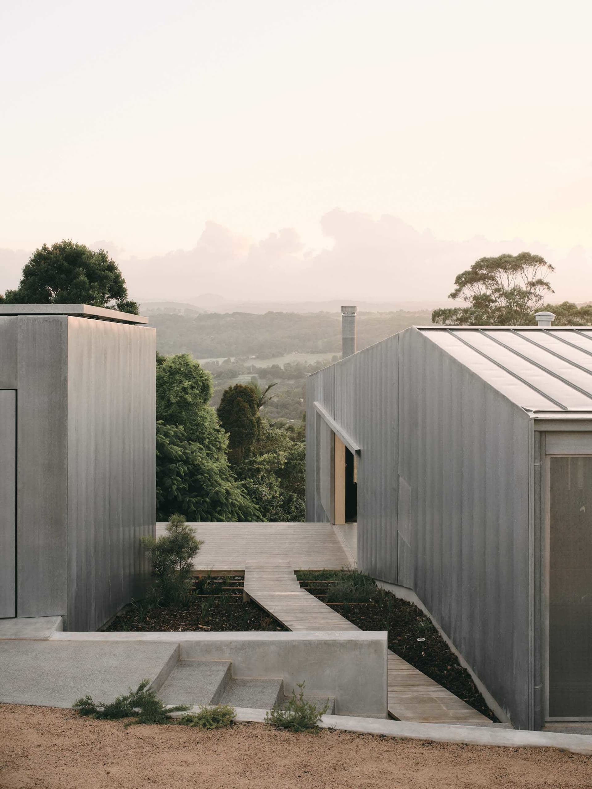 Modern house clad in weathered Corten steel with wooden deck and stairs, set against a vast forested mountain valley at dusk.