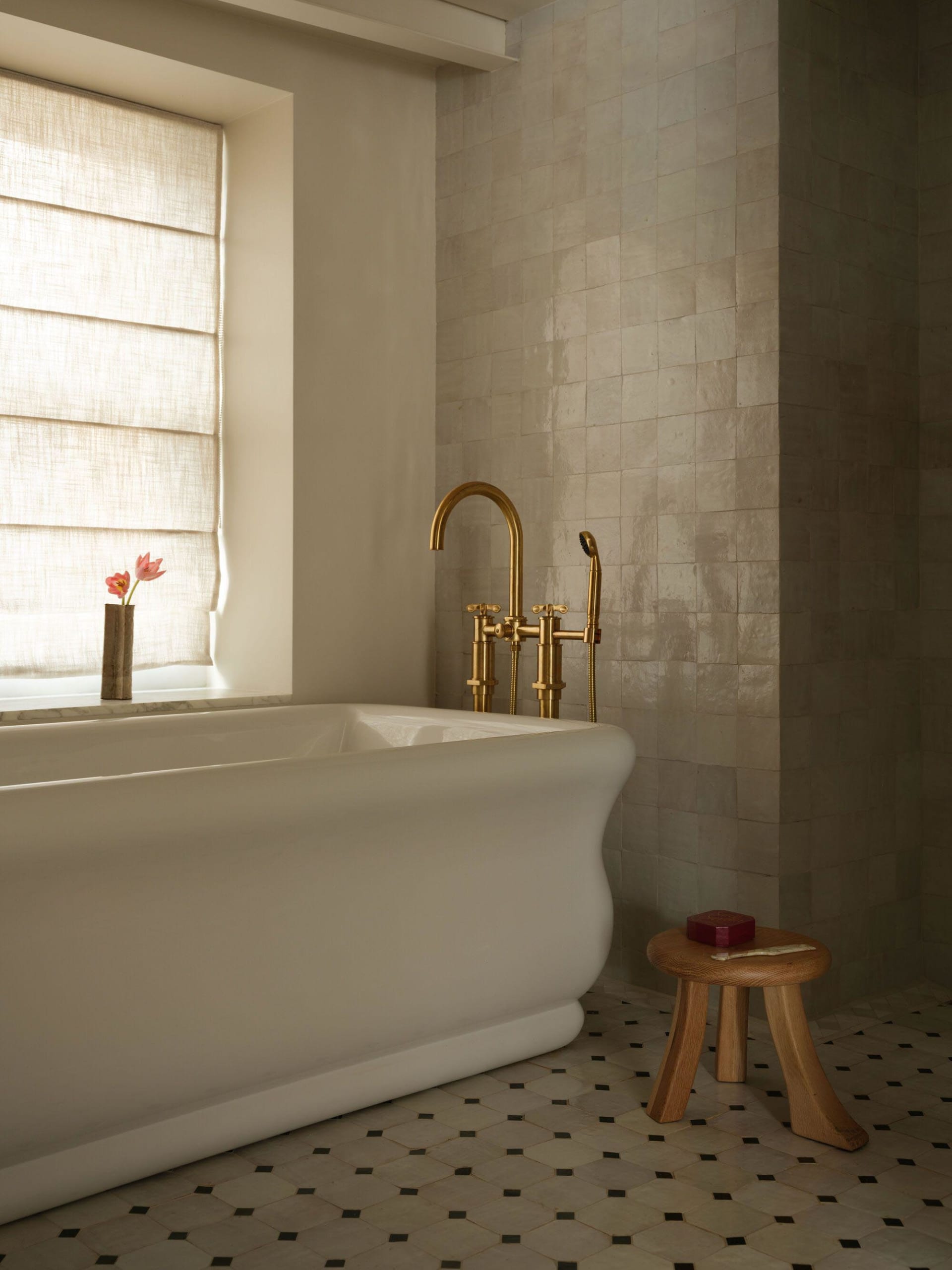 A serene bathroom corner featuring a white freestanding bathtub, unlacquered brass faucet, wooden stool, and beige zellige tiled walls with a window dressed in natural linen roman shades.