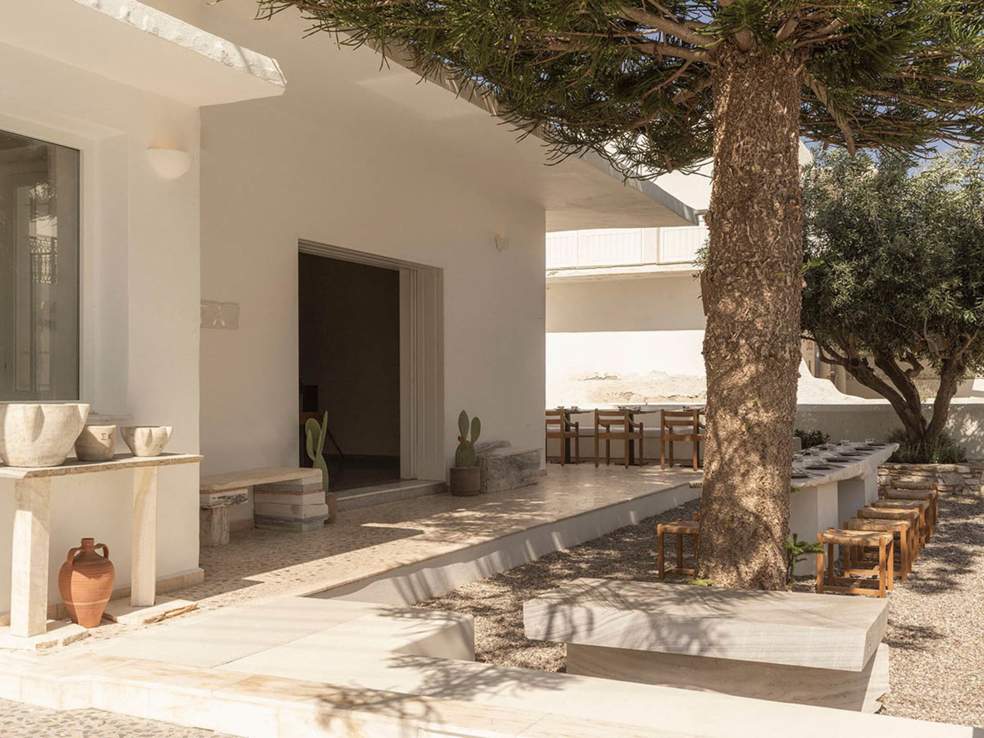 Bright Mediterranean courtyard with white plastered walls, travertine stone benches and tables, a large olive tree providing shade, and terracotta pottery on a stone console.