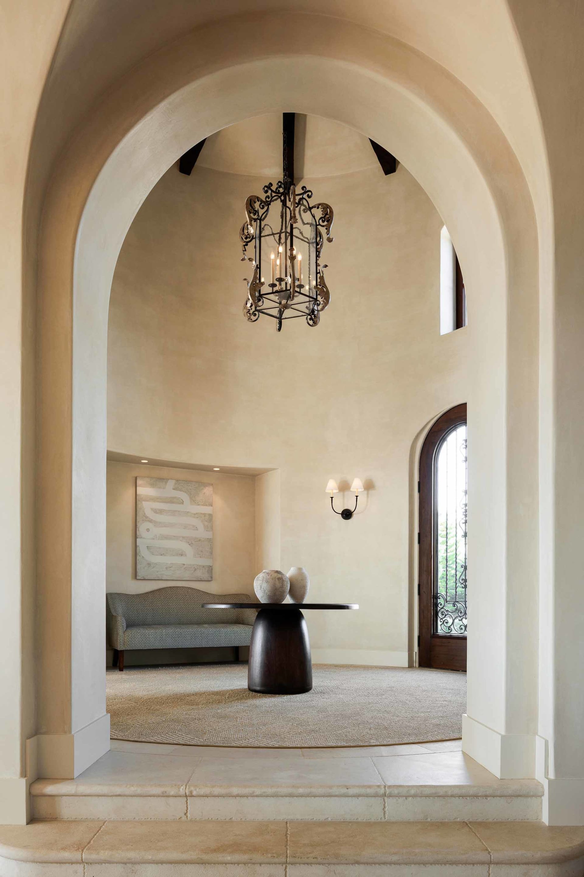 View through a tall arched doorway into a spacious entry hall featuring a dramatic black iron chandelier, vaulted plaster ceilings, a round dark wood pedestal table with ceramic vases, and a patterned settee beneath abstract wall art.