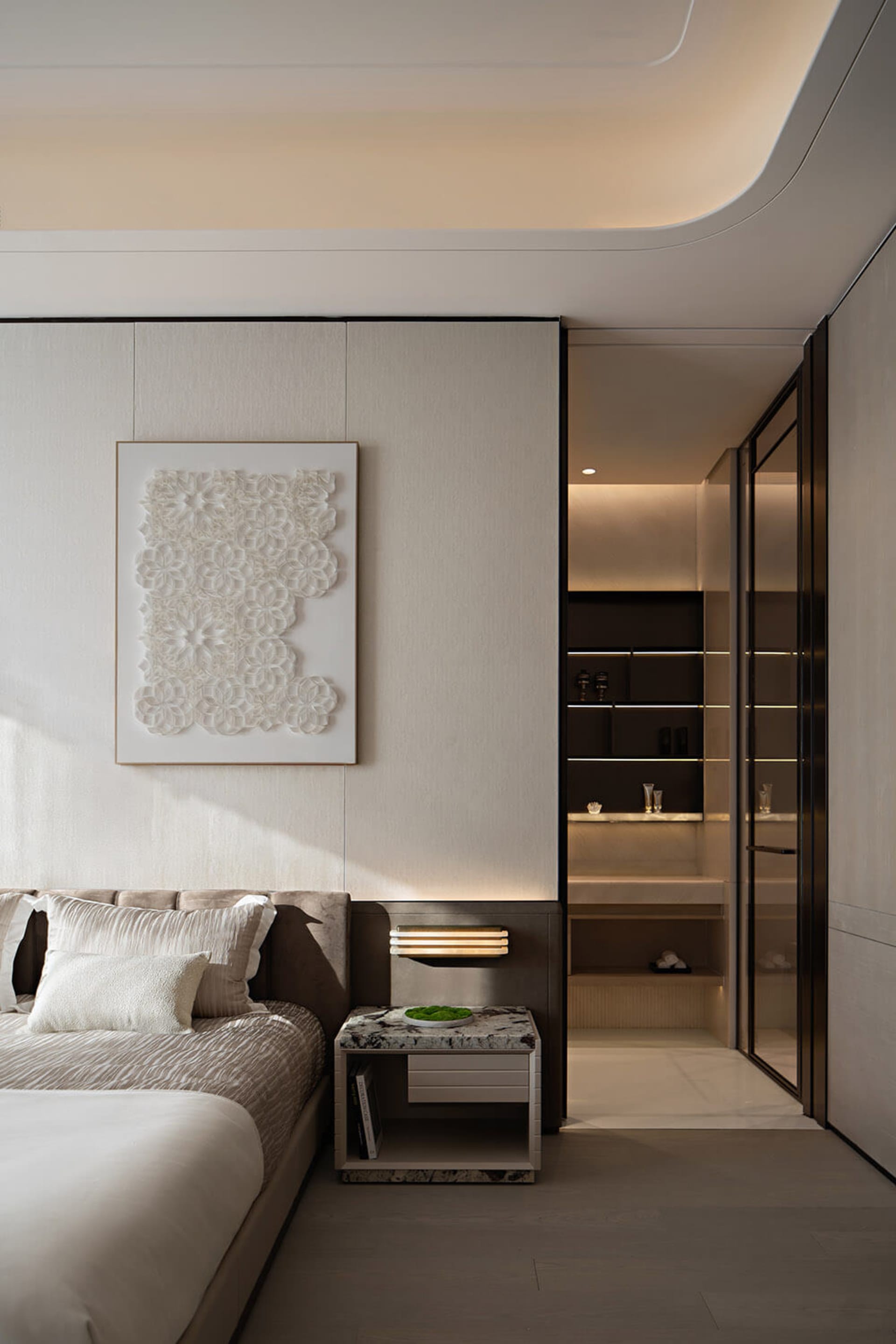 Modern minimalist bedroom featuring a beige upholstered bed with white pillows, a black and white marble nightstand with integrated warm lighting, and a large framed 3D white floral artwork on a textured wall.