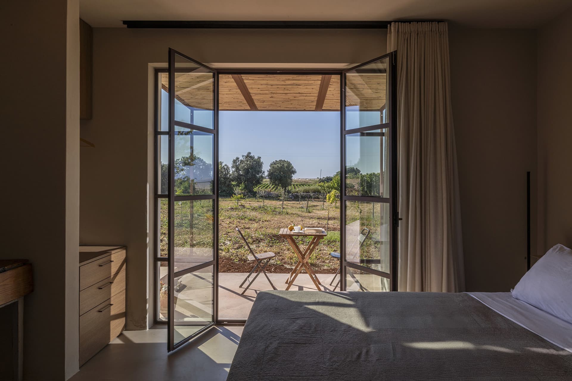 Modern minimalist bedroom featuring a large bed with grey linen, light wood dresser, and wide open black-framed bifold glass doors revealing a vineyard terrace and rural landscape beyond.
