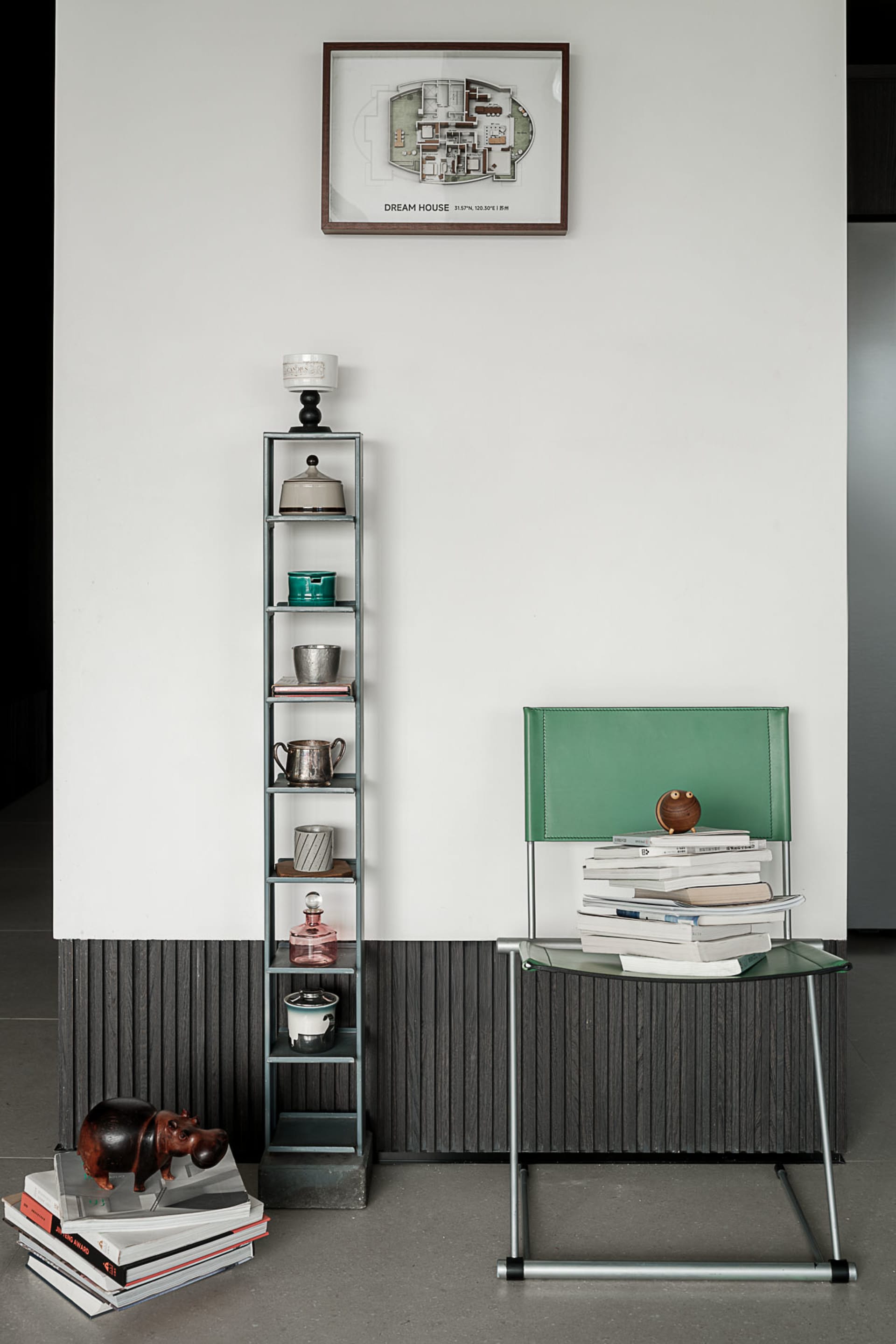 Modern minimalist entryway featuring a tall grey metal shelving unit displaying vintage objects, a bright green leather chair with stacked books, and a framed architectural floor plan labeled 'DREAM HOUSE' on a white wall with dark wood wainscoting.