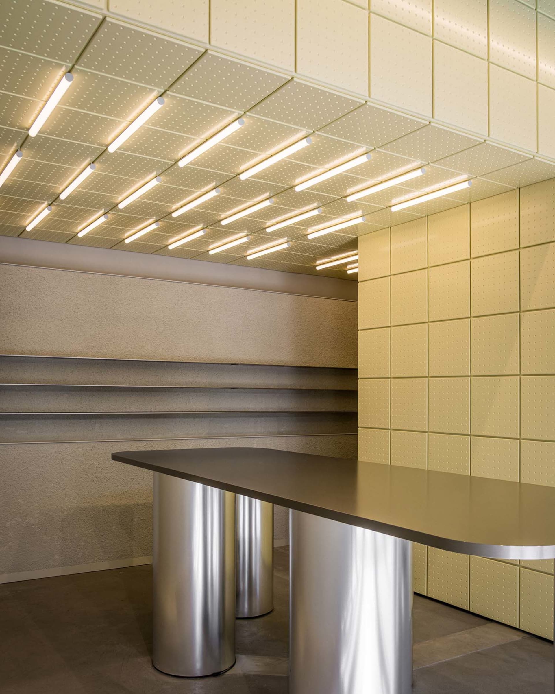 Modern reception desk featuring a curved dark countertop supported by cylindrical stainless steel bases, set against pale yellow perforated wall and ceiling panels with integrated linear lighting.