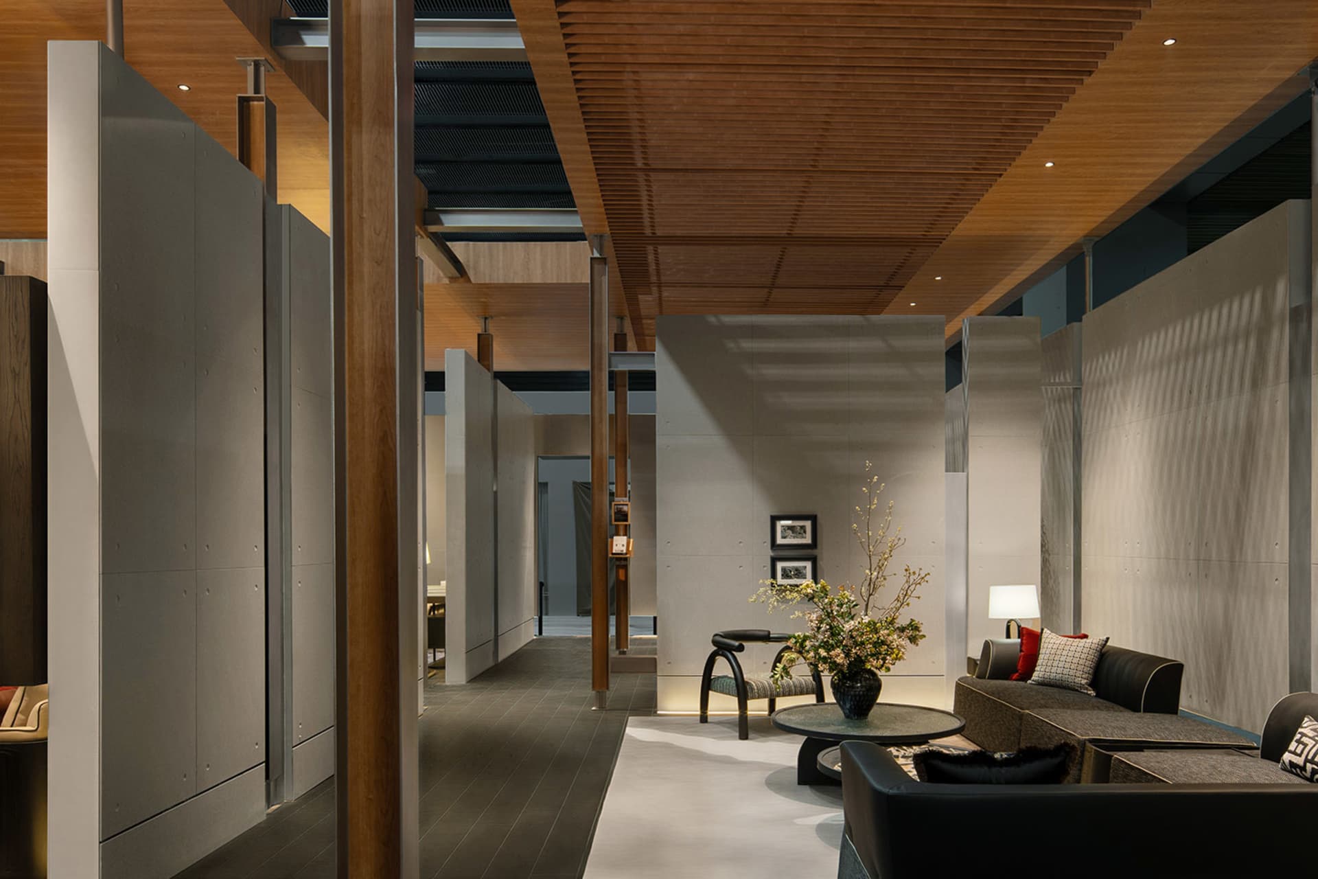 Modern open-plan living area featuring concrete walls, wooden ceiling slats, a dark sectional sofa, and a large floral arrangement on a round coffee table.