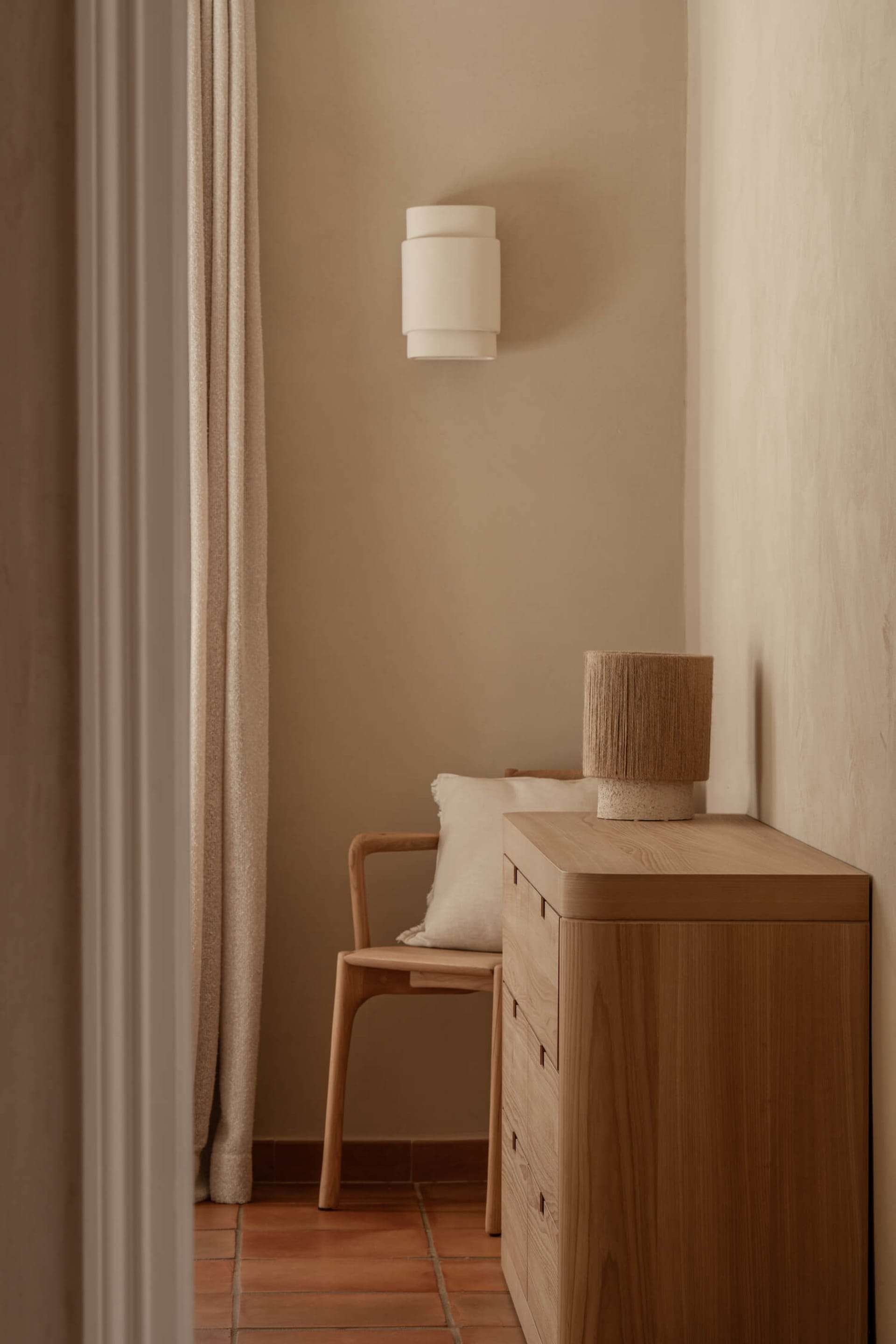 Minimalist entryway featuring light oak dresser, wooden armchair with linen cushion, cylindrical wall sconce, and terracotta tile floor in warm neutral tones.