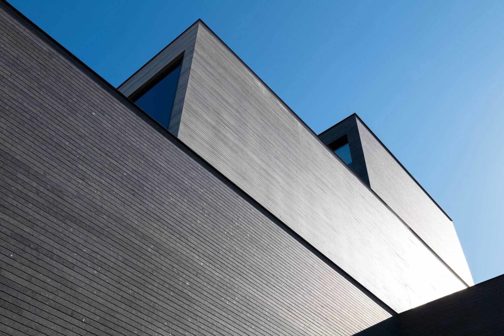 Low-angle view of a contemporary building with dark horizontal wood cladding and distinctive triangular windows set against a clear blue sky.