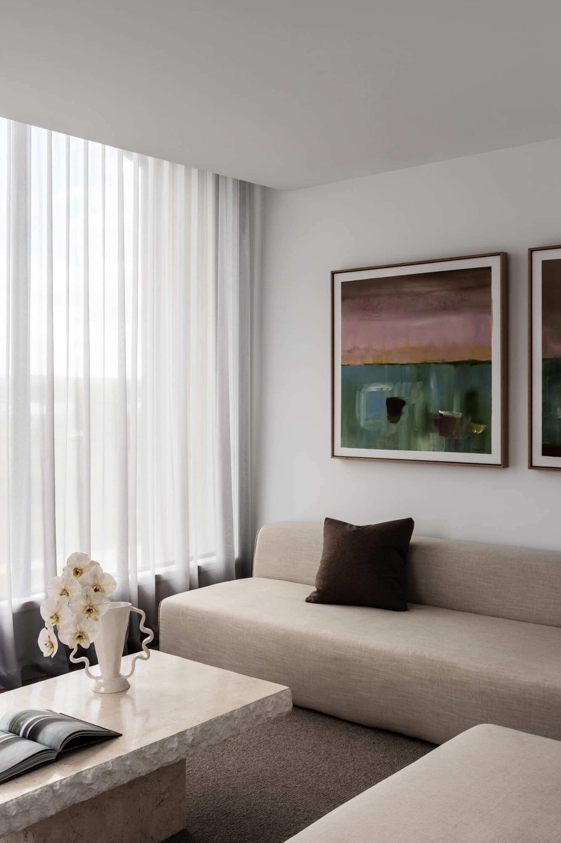 Modern minimalist living room featuring a beige sofa with brown cushion, a large travertine coffee table with white orchid arrangement, and two abstract landscape paintings on a white wall.