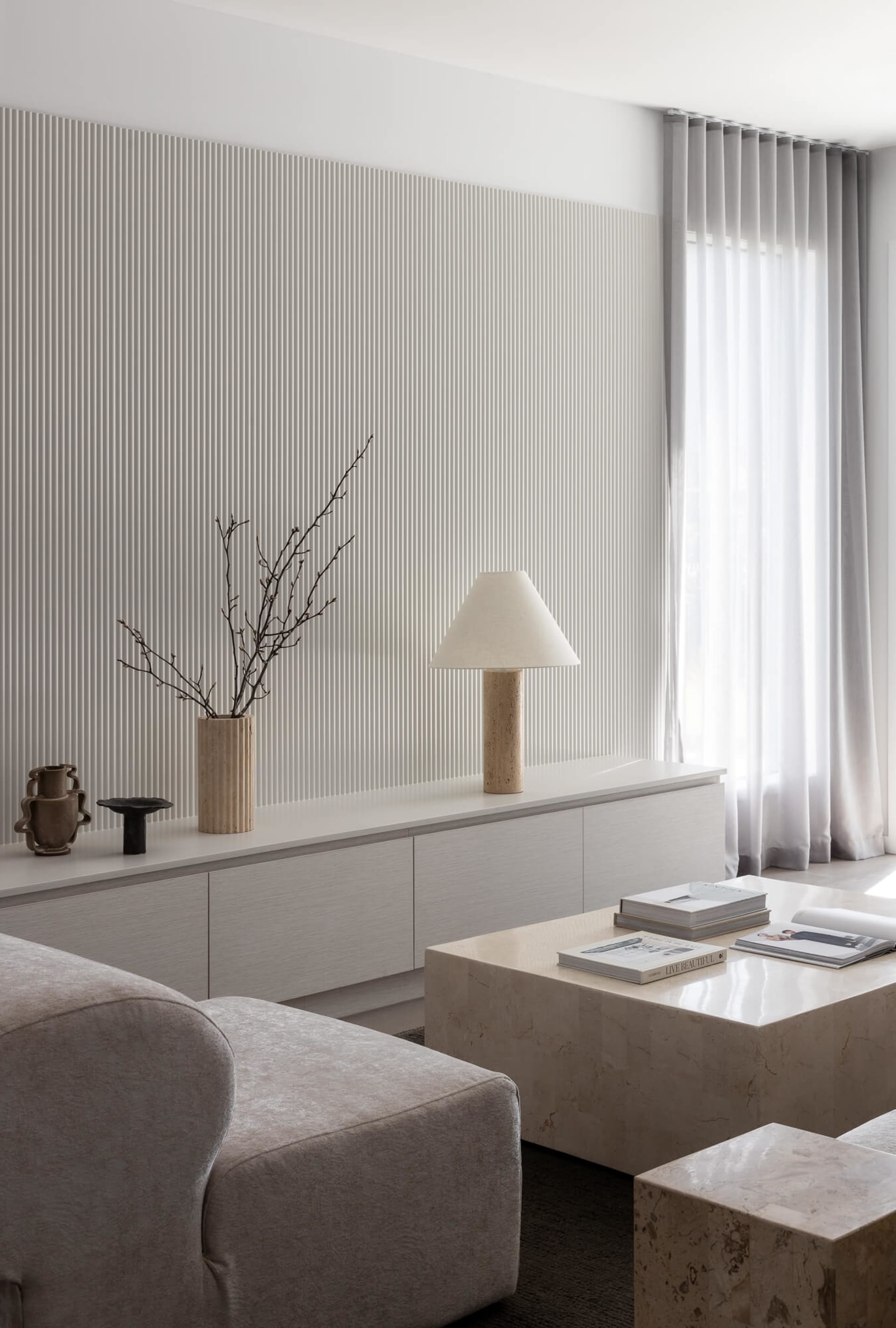 Modern minimalist living room featuring a tall fluted beige accent wall, low white sideboard with travertine lamp and dried branches, and large travertine coffee tables with books, paired with a soft beige bouclé sofa.