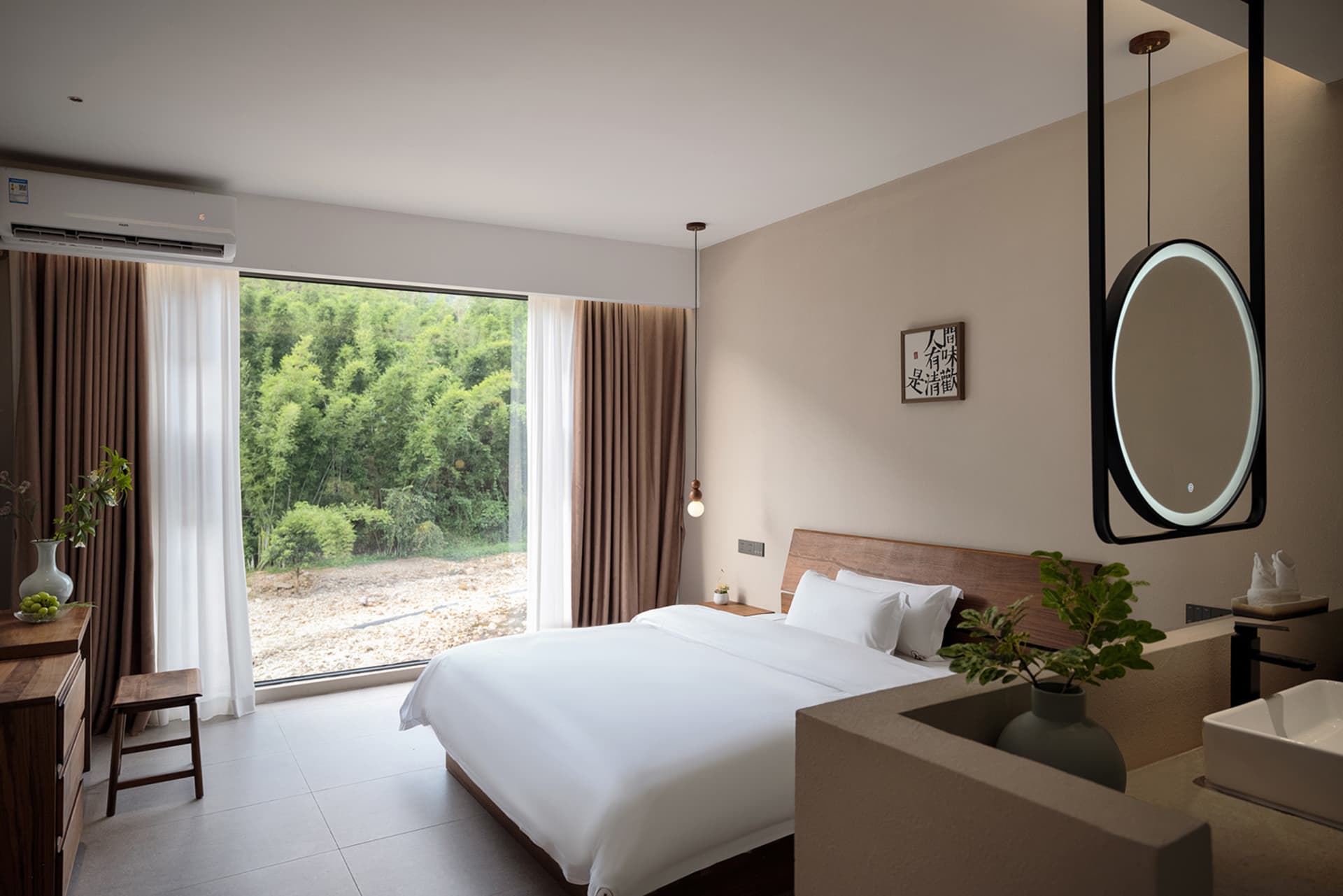 Modern hotel bedroom featuring a walnut wood headboard, white bedding, large glass doors opening to a bamboo forest, pendant light, and integrated vanity with round backlit mirror.
