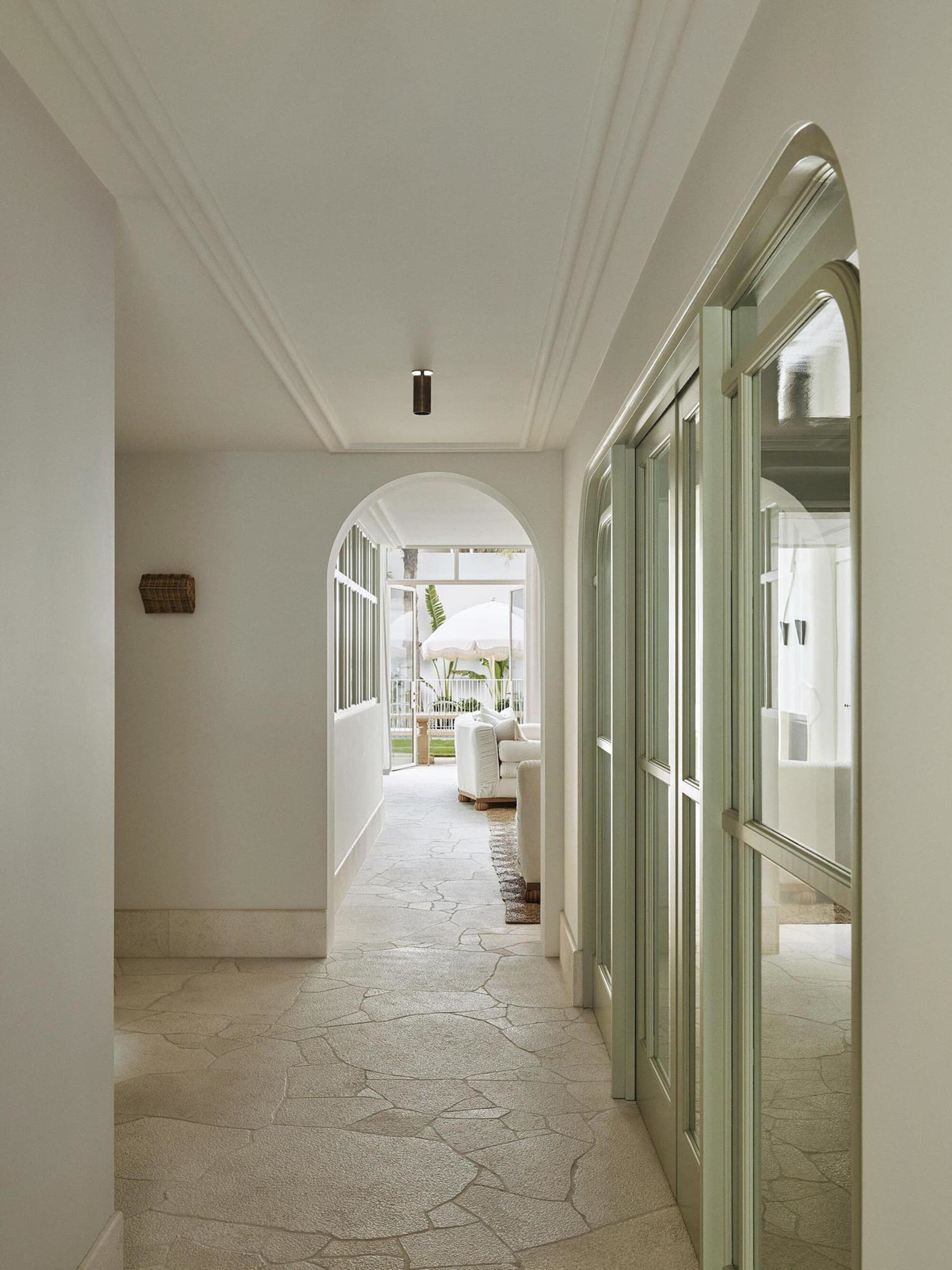 Long white hallway with arched doorways, pale limestone flagstone floor, and views to an outdoor patio with white furniture.