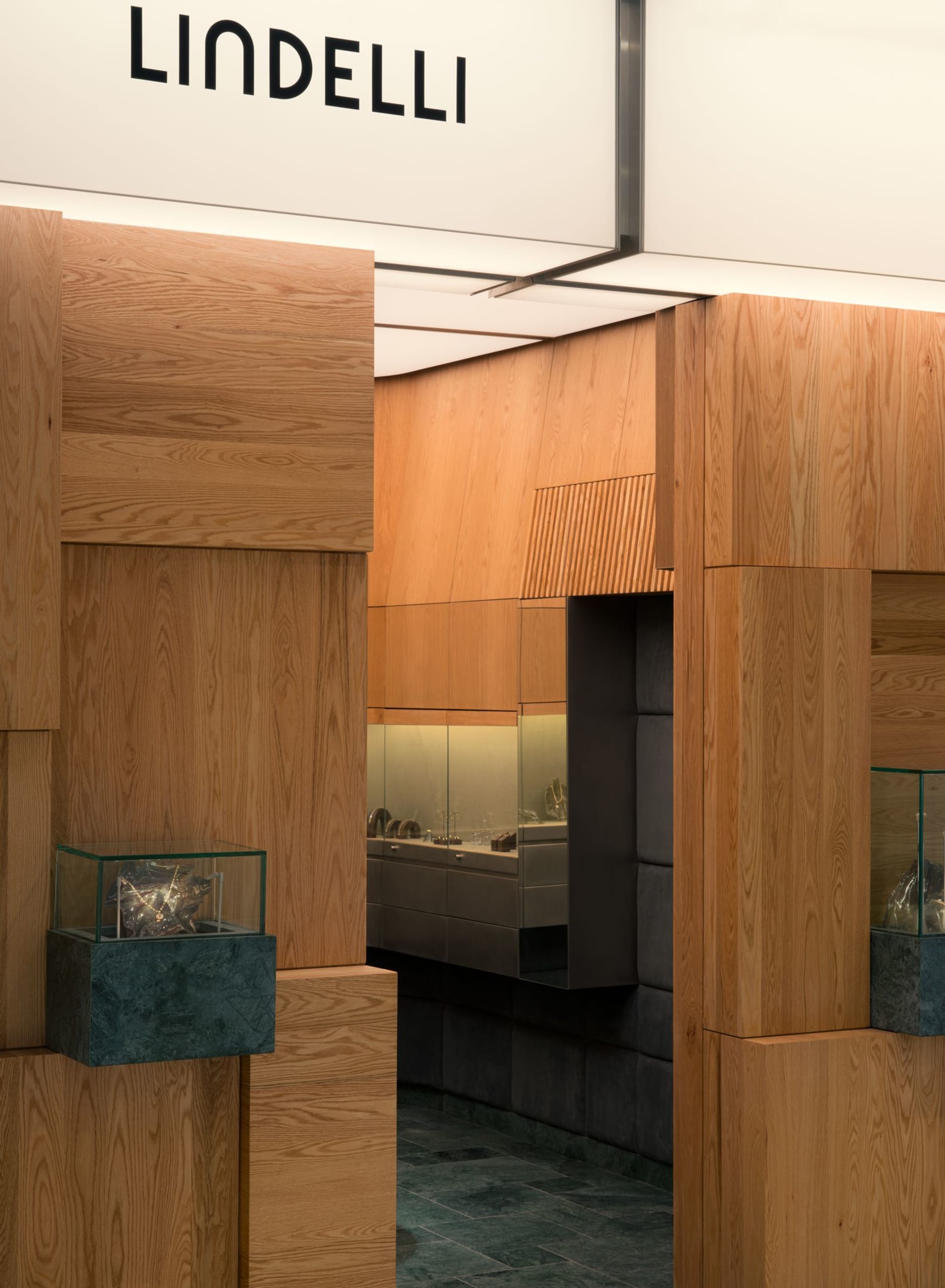Modern jewelry boutique interior featuring light oak wood paneling, dark green marble bases, glass display cases, and integrated lighting.