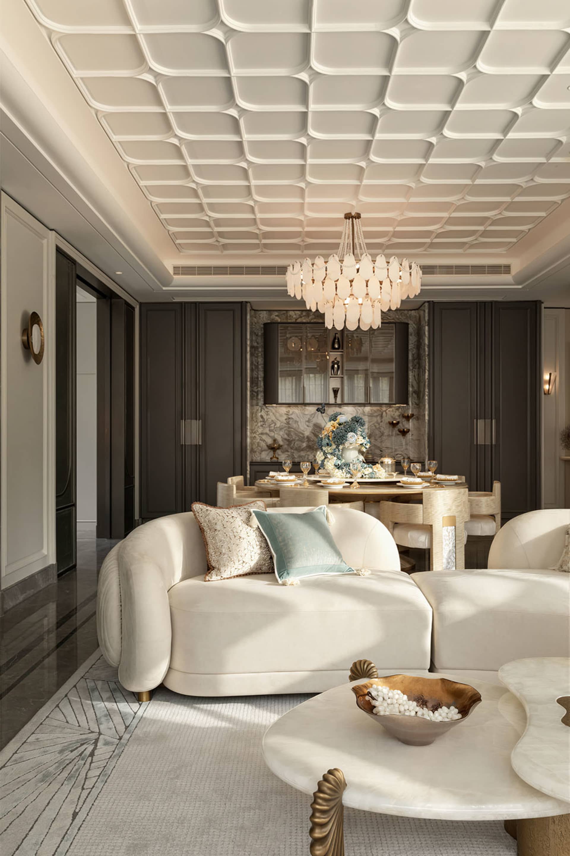 Modern luxury living and dining area featuring a white curved sofa with decorative pillows, marble coffee tables, a large shell chandelier, and a dramatic 3D geometric ceiling in neutral tones.