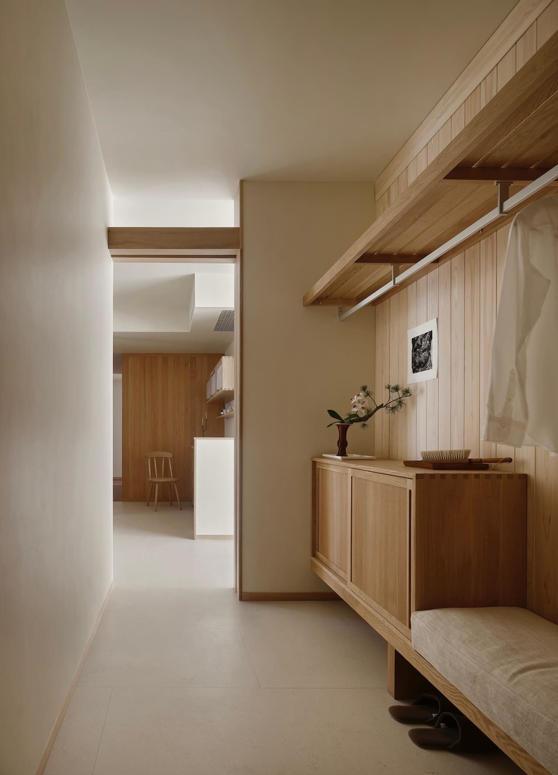Modern minimalist entryway with light oak built-in bench, upper hanging rod, cabinet storage, and a view into a bright kitchen area.