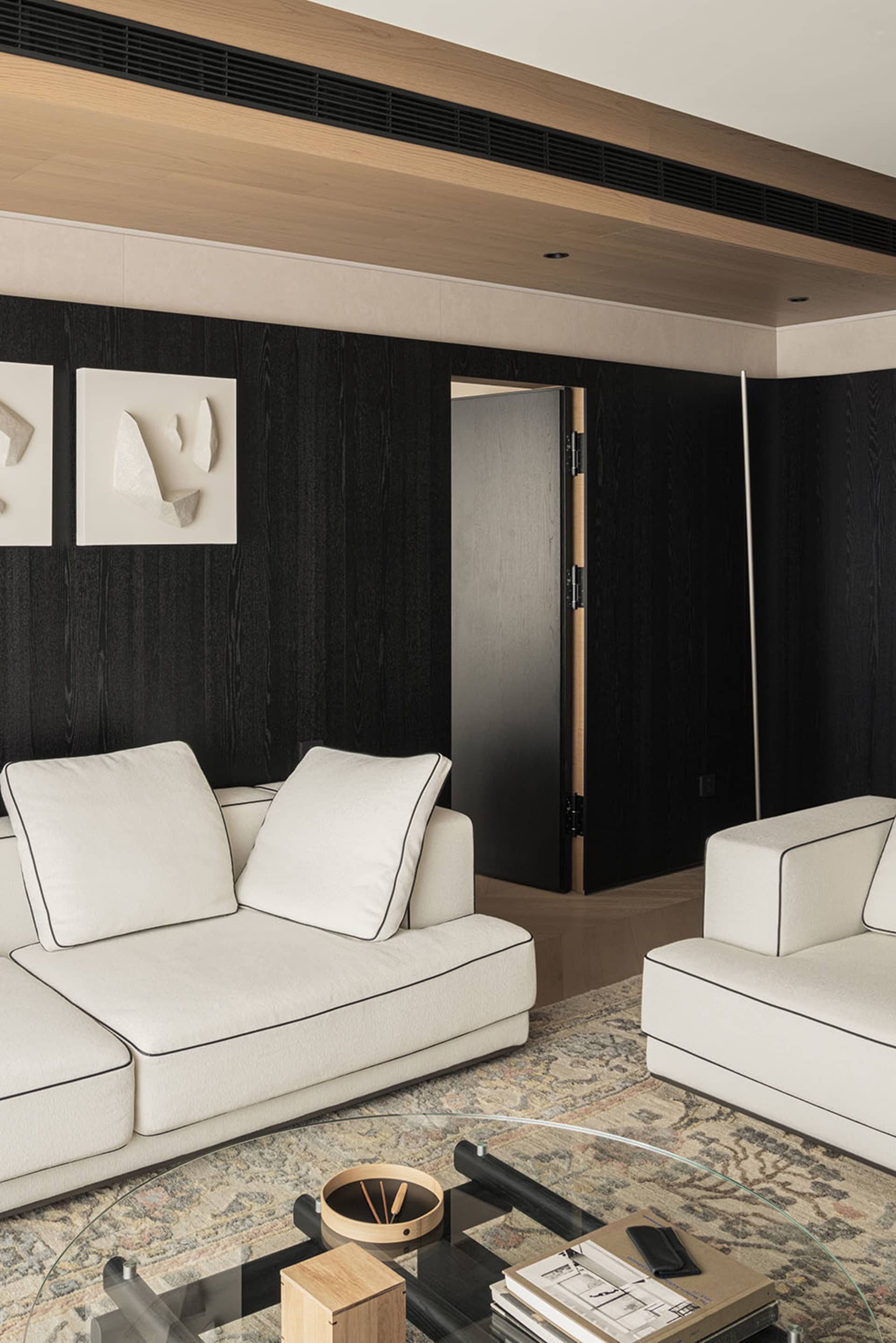 Modern living room featuring a black oak accent wall, white sofas with black piping, abstract wall art, and a patterned area rug with a round glass coffee table.