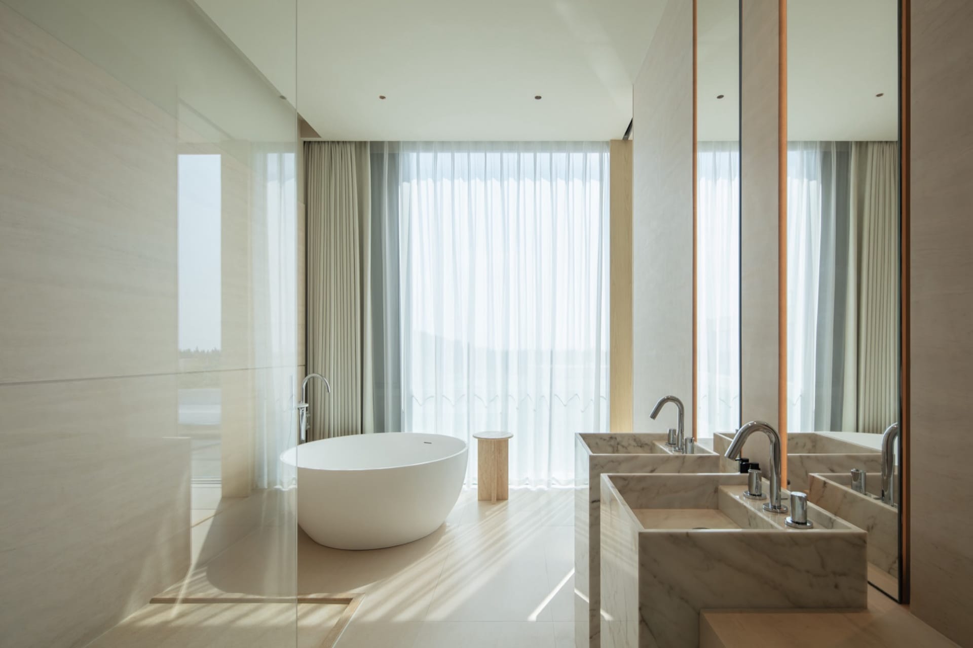 Modern minimalist bathroom featuring a white freestanding oval bathtub, double marble rectangular sinks with chrome faucets, large mirrors, sheer curtains, and abundant natural light.