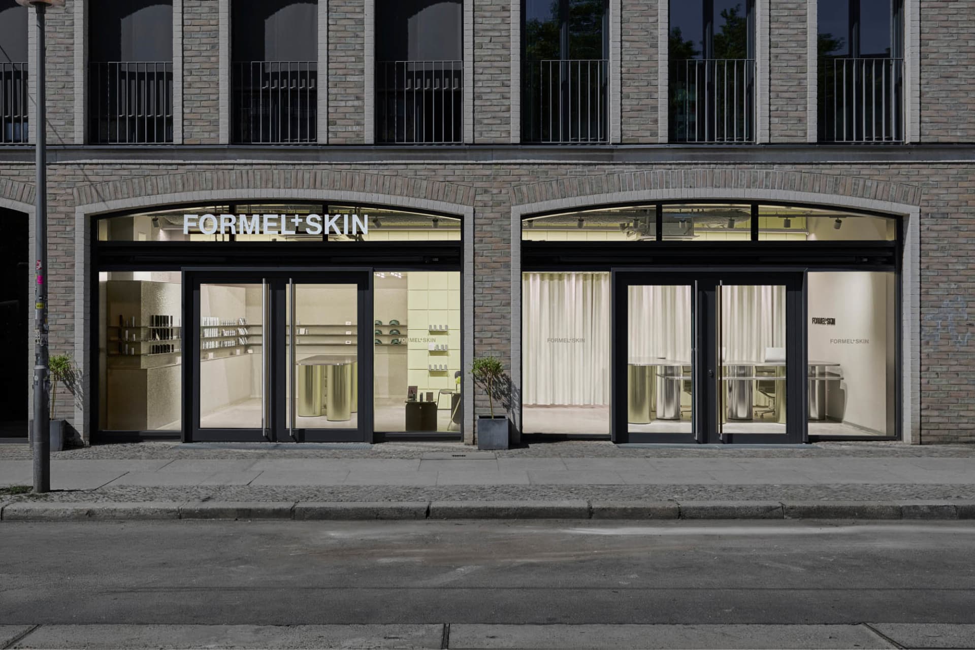Street view of Formel Skin beauty clinic storefront in Berlin, showcasing two arched glass entrances with black frames, cylindrical brass display tables, pale yellow interior walls, and potted trees on the sidewalk.