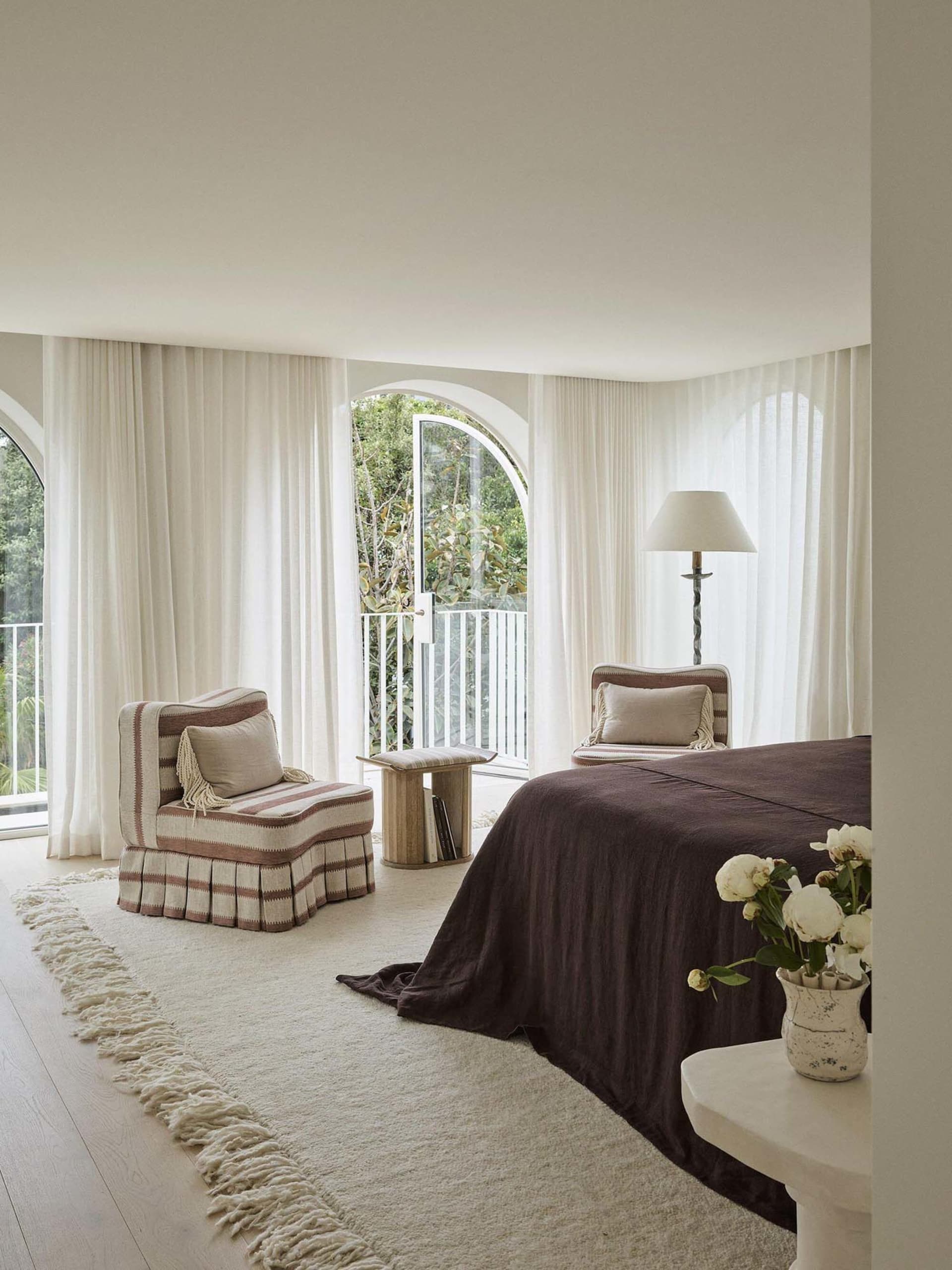 Bright modern bedroom featuring arched windows with sheer curtains, two striped linen armchairs, a large bed with chocolate brown linen coverlet, and a cream shaggy rug on light wood flooring.